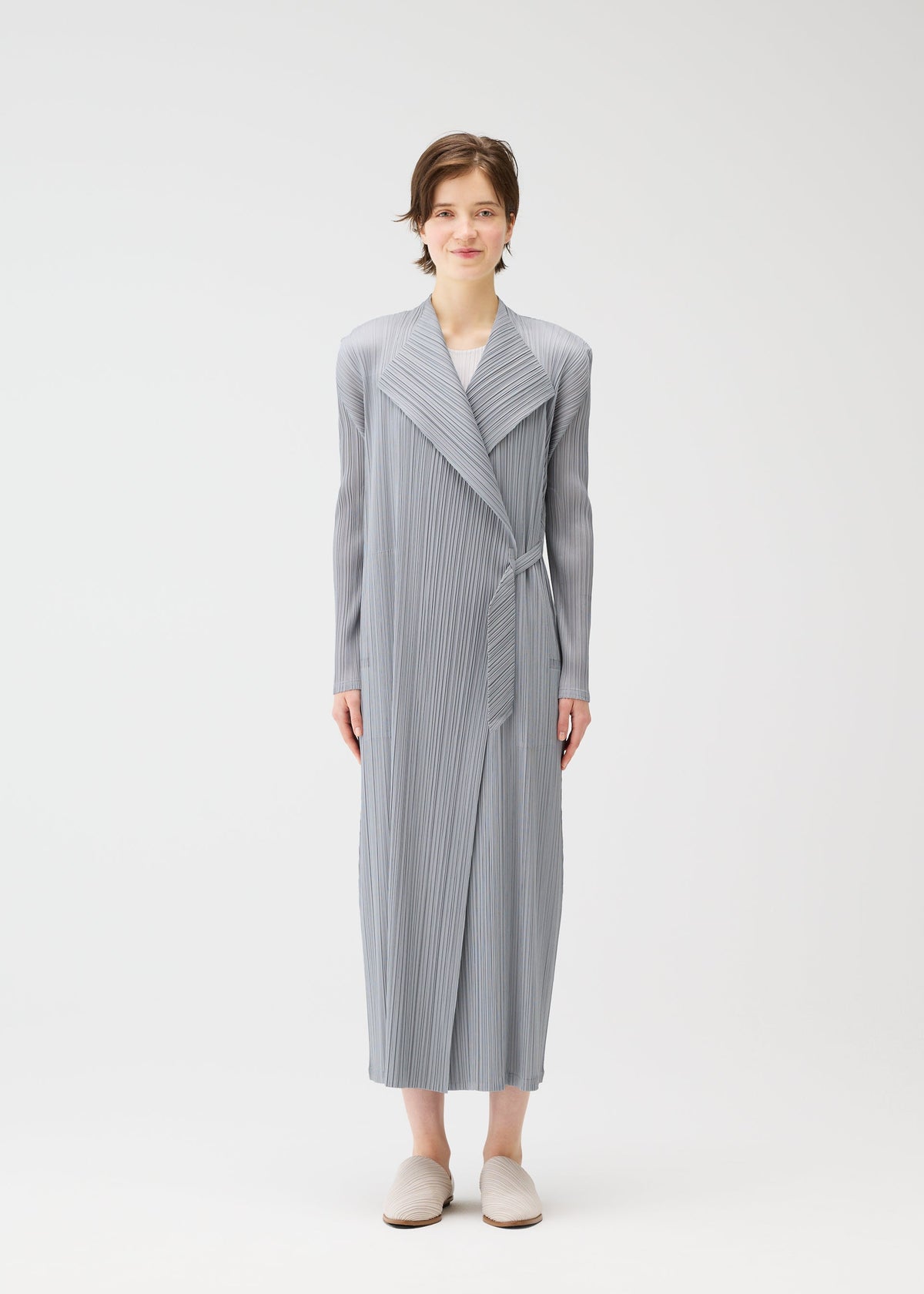A person faces forward, wearing the FACADE long coat by PLEATS PLEASE ISSEY MIYAKE with a light gray pleated wrap and subtle cord details, paired with beige slippers, against a plain white background. view 6 of 9
