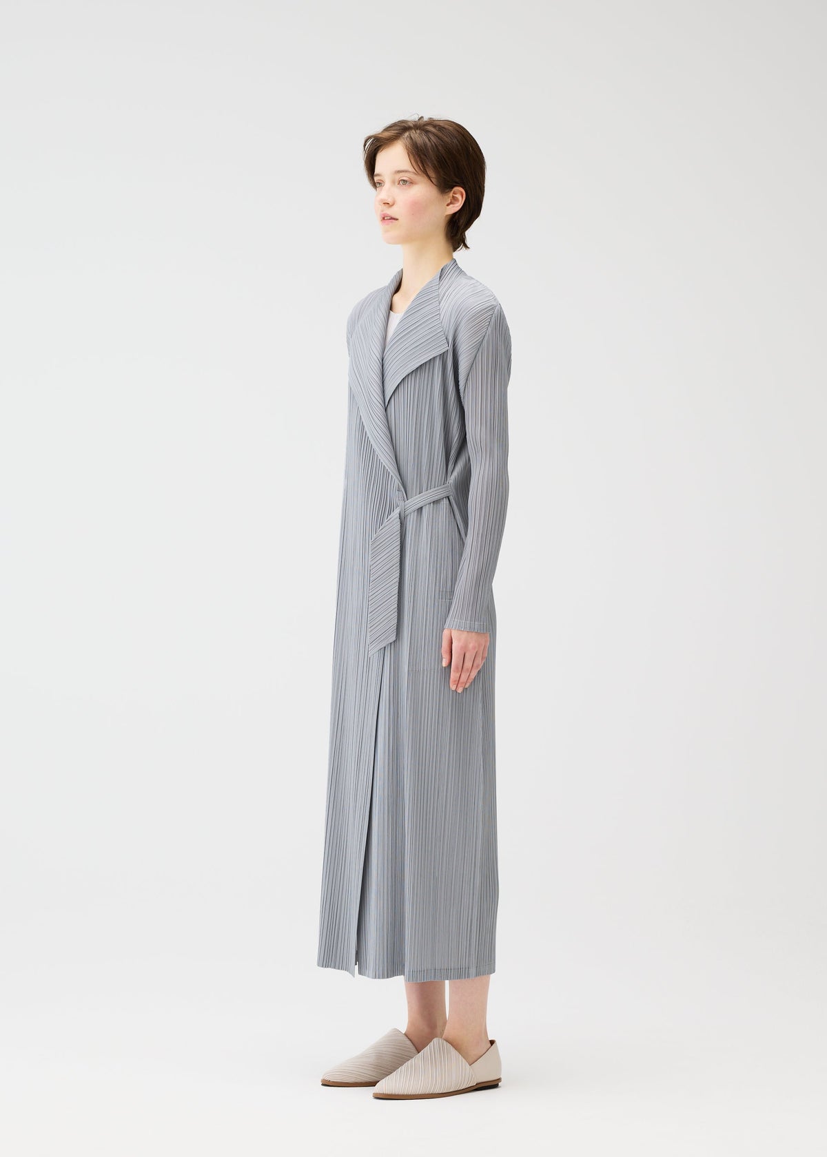 A person stands sideways in the FACADE robe by PLEATS PLEASE ISSEY MIYAKE—long, light gray, pinstriped with a cord belt and matching slippers—against a plain white background. view 7 of 9