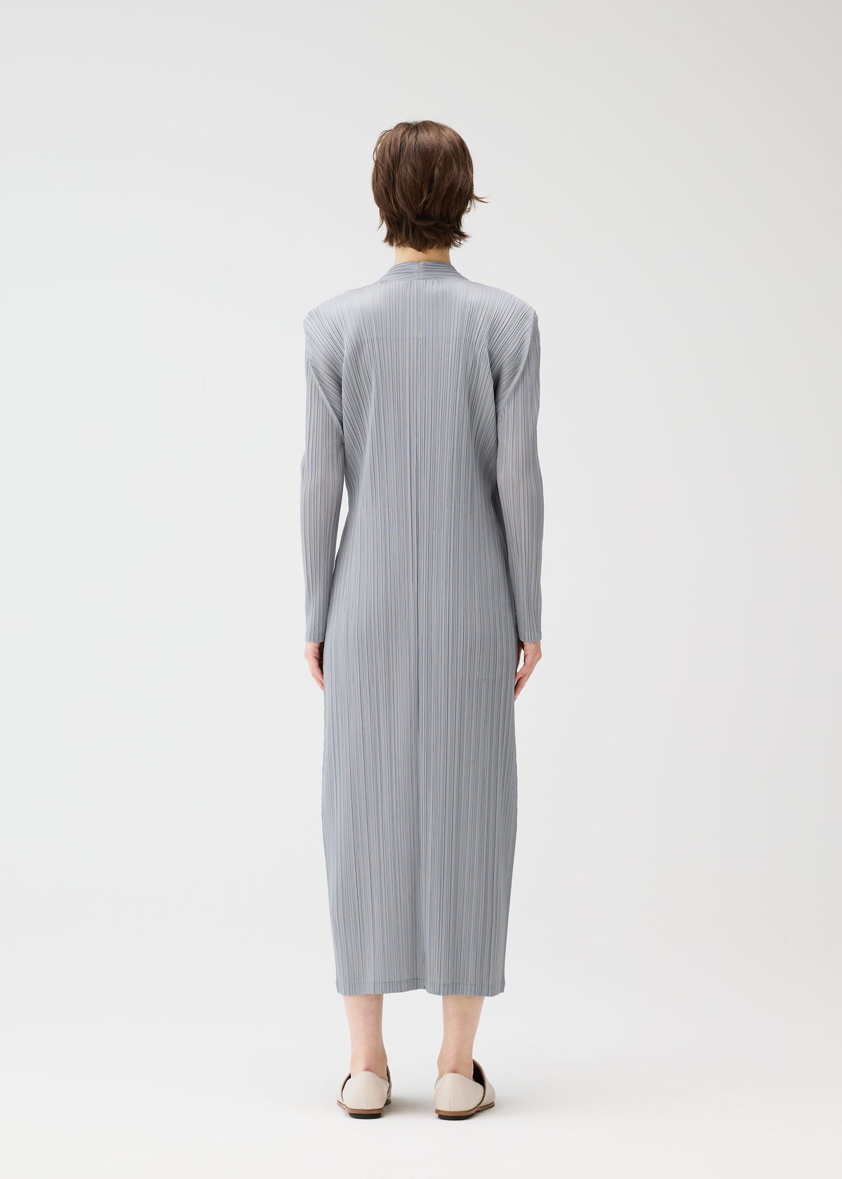 A person with short brown hair faces away, wearing the FACADE long, light gray pleated dress from PLEATS PLEASE ISSEY MIYAKE under a subtly corded long coat, with beige flats, against a plain white background. view 8 of 9