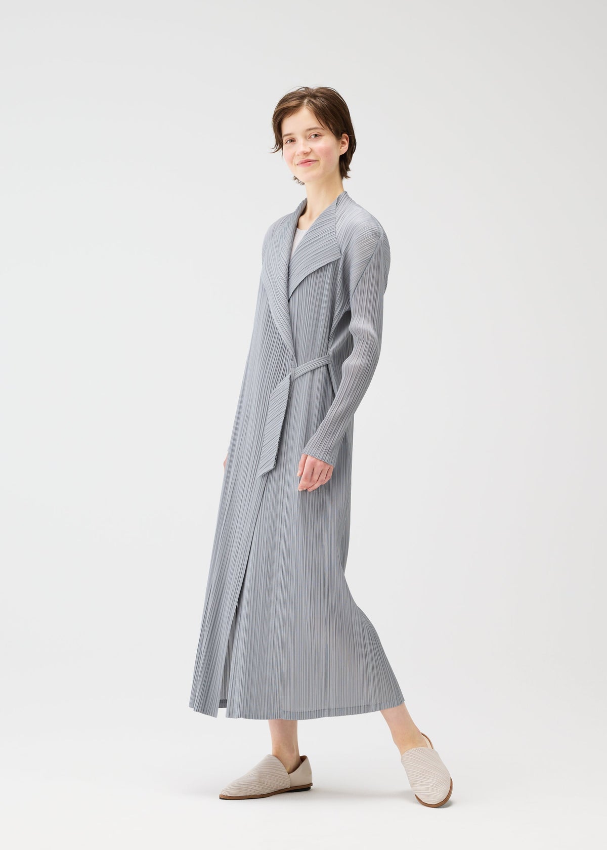A person wears the FACADE pleated robe by PLEATS PLEASE ISSEY MIYAKE with a cord-design belt and matching slip-ons, standing against a white background. They have short hair, a slight smile, and one foot raised in mid-step. view 9 of 9