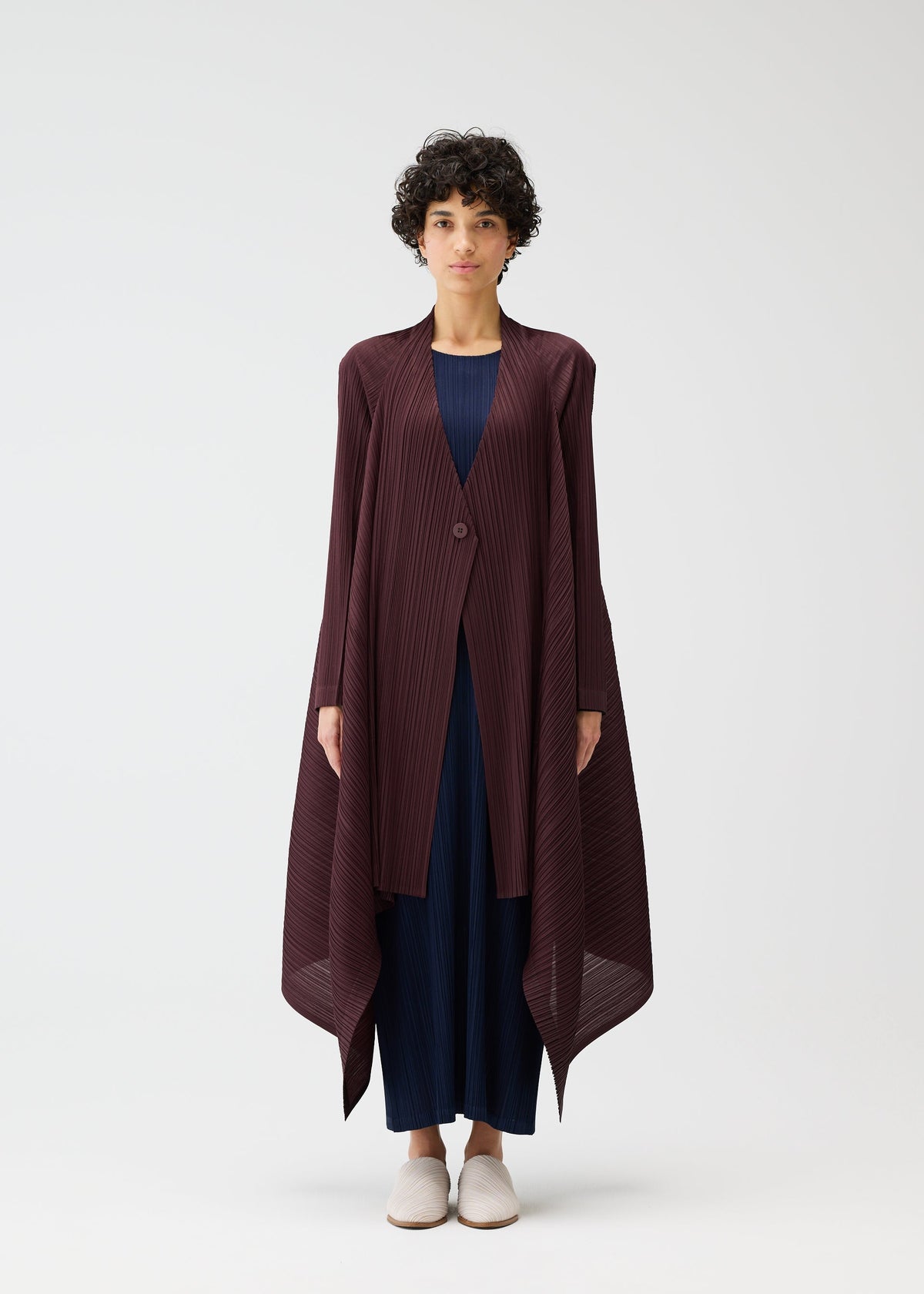 A person with short curly hair stands against a plain background, wearing the CITY FRAME long pleated cardigan in dark burgundy from PLEATS PLEASE ISSEY MIYAKE, styled over a navy outfit and light shoes. The jacket has an asymmetrical hem. view 5 of 6