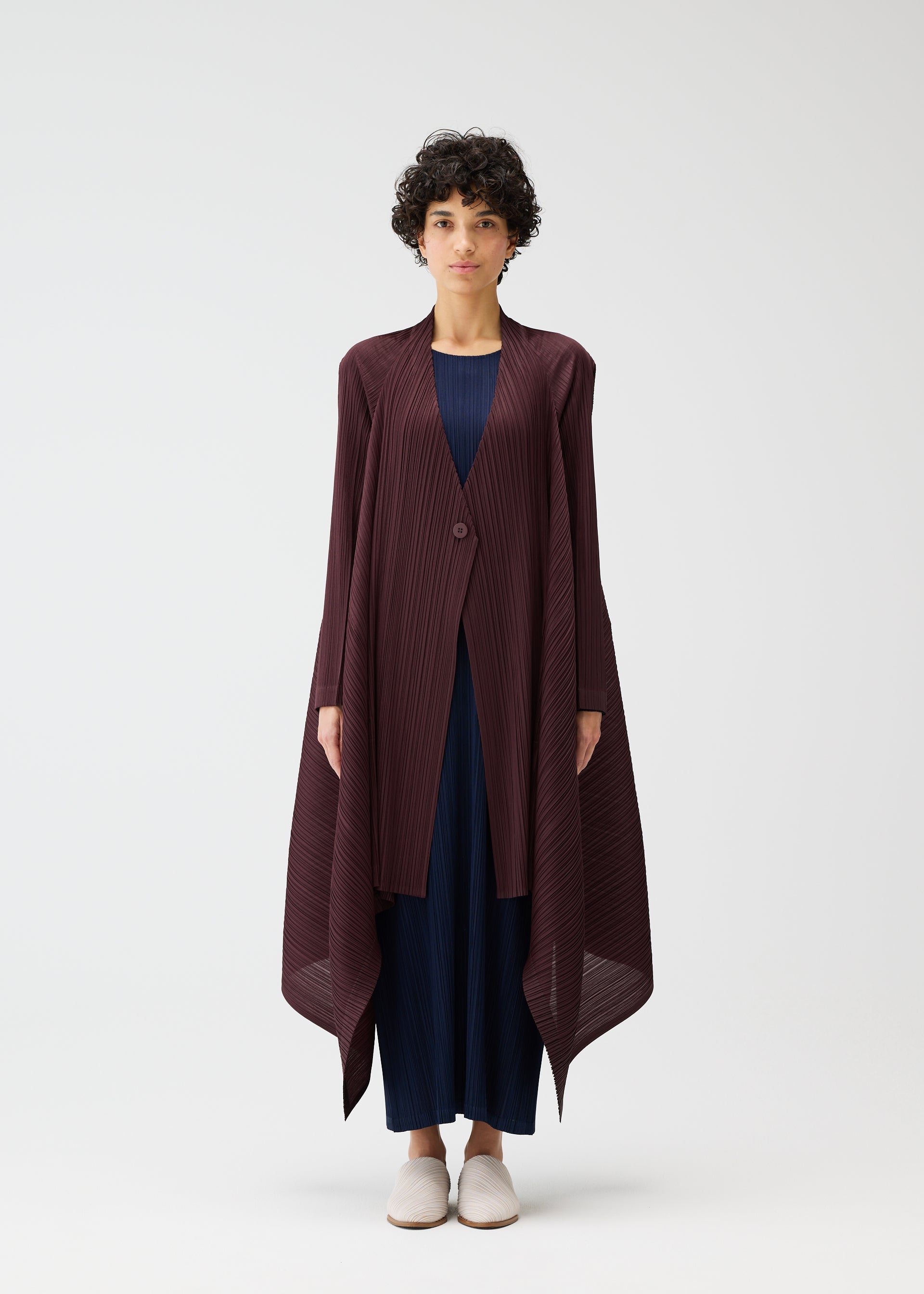 Zoomed view A person with short curly hair stands against a plain background, wearing the CITY FRAME long pleated cardigan in dark burgundy from PLEATS PLEASE ISSEY MIYAKE, styled over a navy outfit and light shoes. The jacket has an asymmetrical hem.