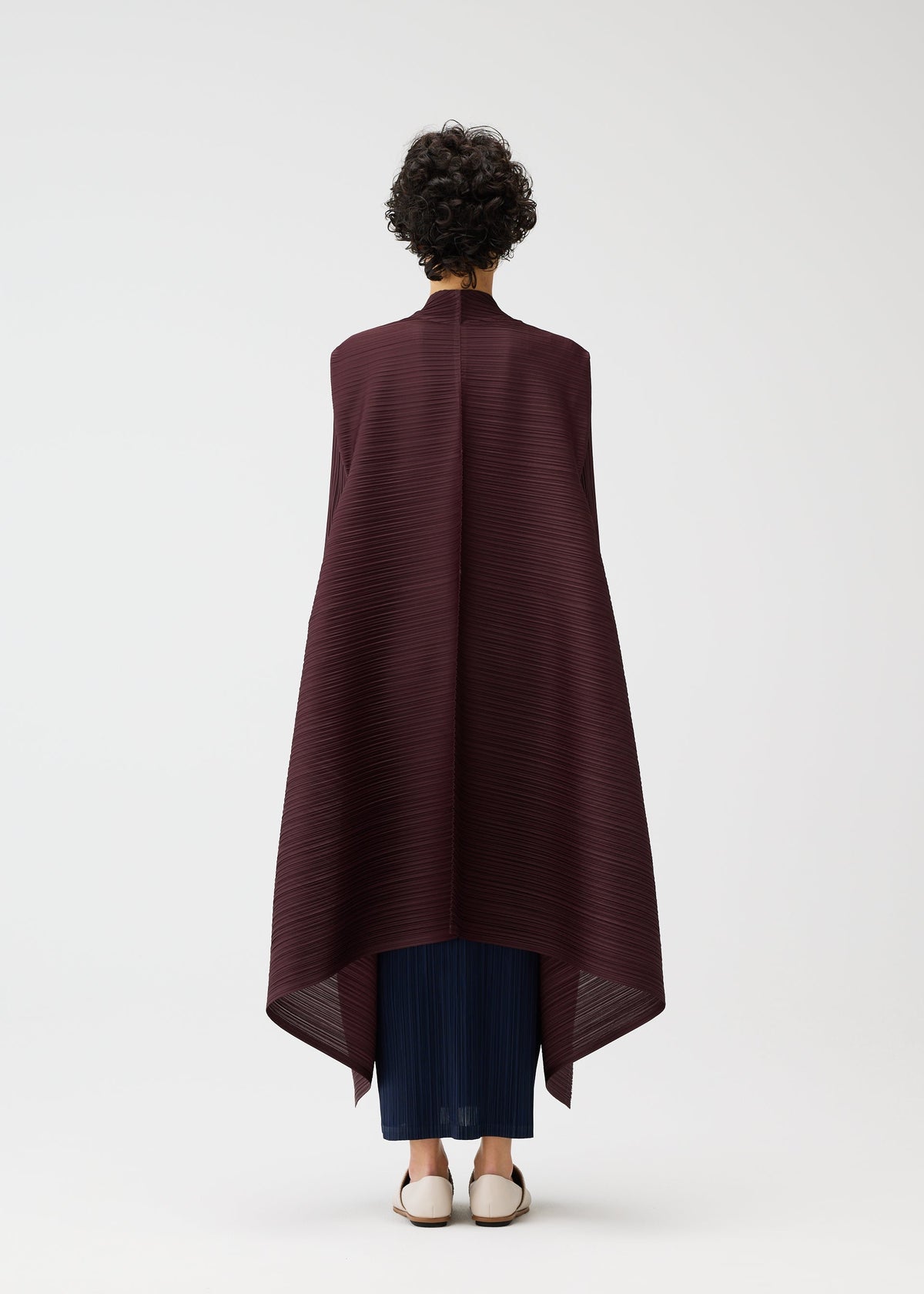 A person with short curly hair faces away, wearing the CITY FRAME long dark burgundy pleated cardigan from PLEATS PLEASE ISSEY MIYAKE over dark blue pants and white shoes, set against a plain white background. view 6 of 6