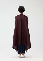 A person with short curly hair faces away, wearing the CITY FRAME long dark burgundy pleated cardigan from PLEATS PLEASE ISSEY MIYAKE over dark blue pants and white shoes, set against a plain white background.