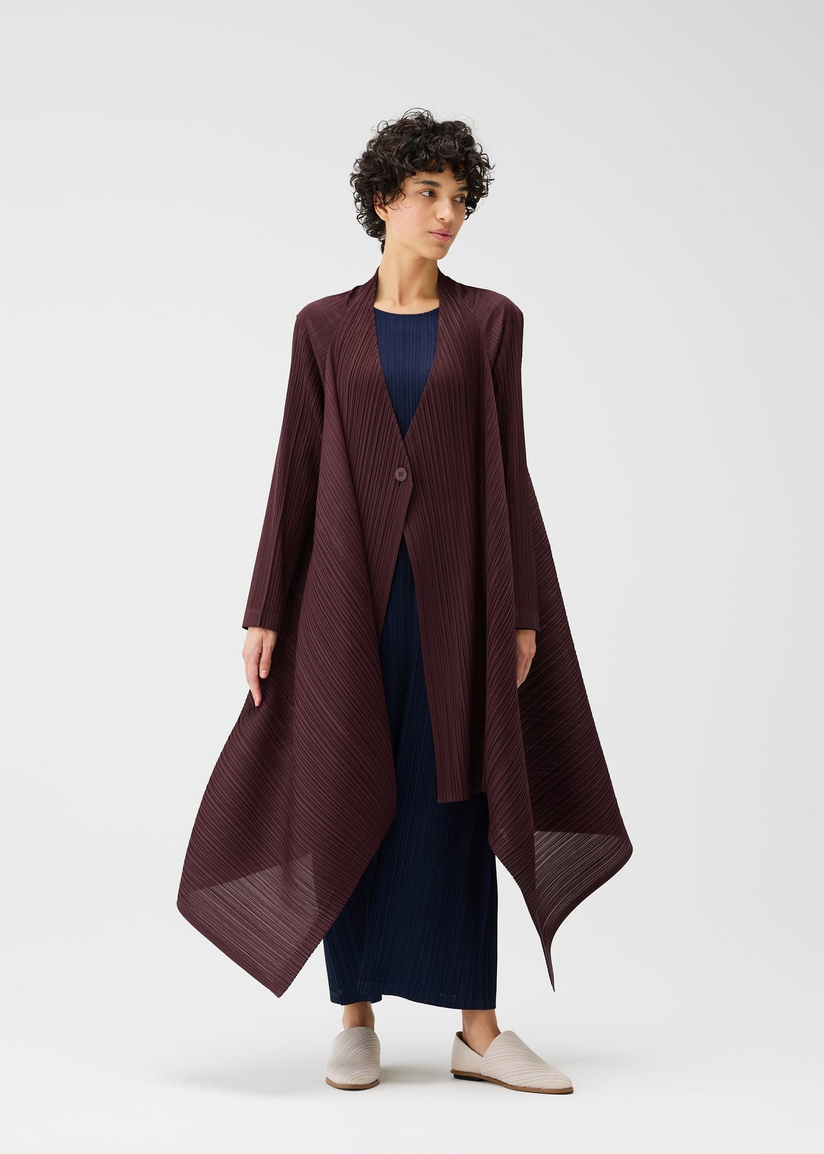 A person with short curly hair models the CITY FRAME voluminous, asymmetrical burgundy pleated coat from PLEATS PLEASE ISSEY MIYAKE over a navy dress and beige shoes, posed against a plain white background. view 4 of 6