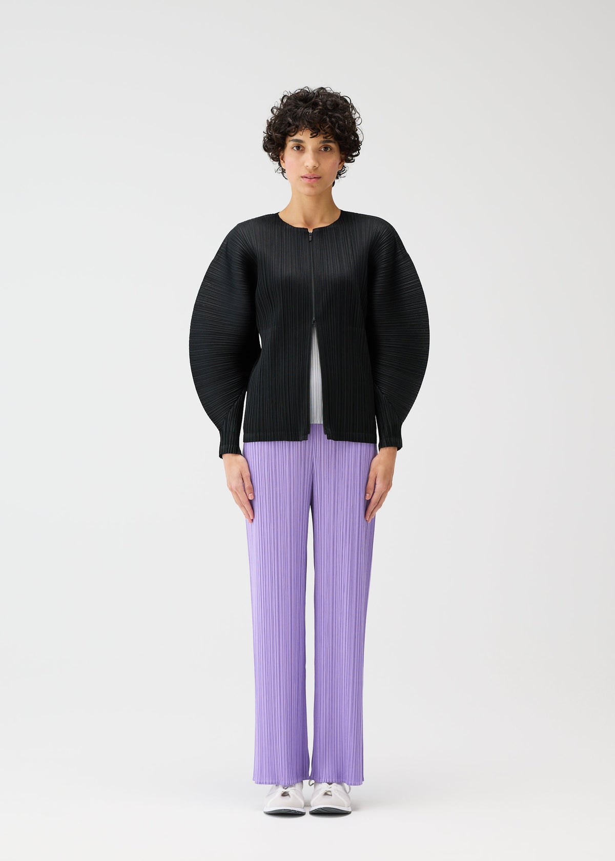 A person faces forward against a white background, wearing the MONTHLY COLORS : APRIL pleated pants by PLEATS PLEASE ISSEY MIYAKE, paired with a black zipper jacket and white shoes. view 8 of 11