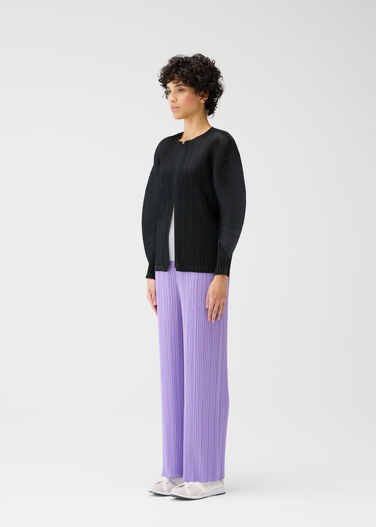A person with short curly hair stands against a plain light background, wearing MONTHLY COLORS : APRIL by PLEATS PLEASE ISSEY MIYAKE—a black long-sleeve top and loose, pleated lavender pants—paired with white shoes. view 9 of 11