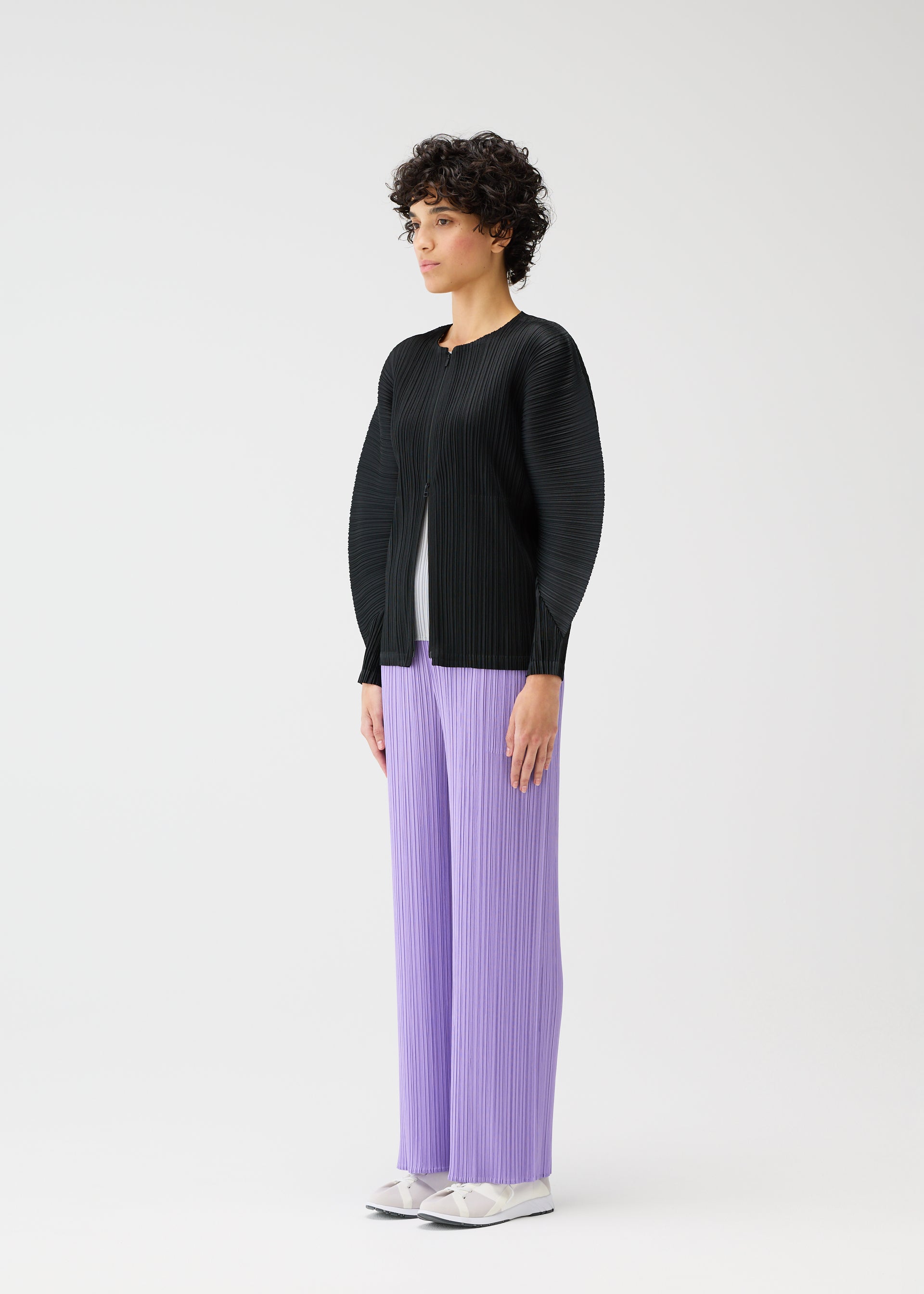 Zoomed view A person with short curly hair stands against a plain light background, wearing MONTHLY COLORS : APRIL by PLEATS PLEASE ISSEY MIYAKE—a black long-sleeve top and loose, pleated lavender pants—paired with white shoes.