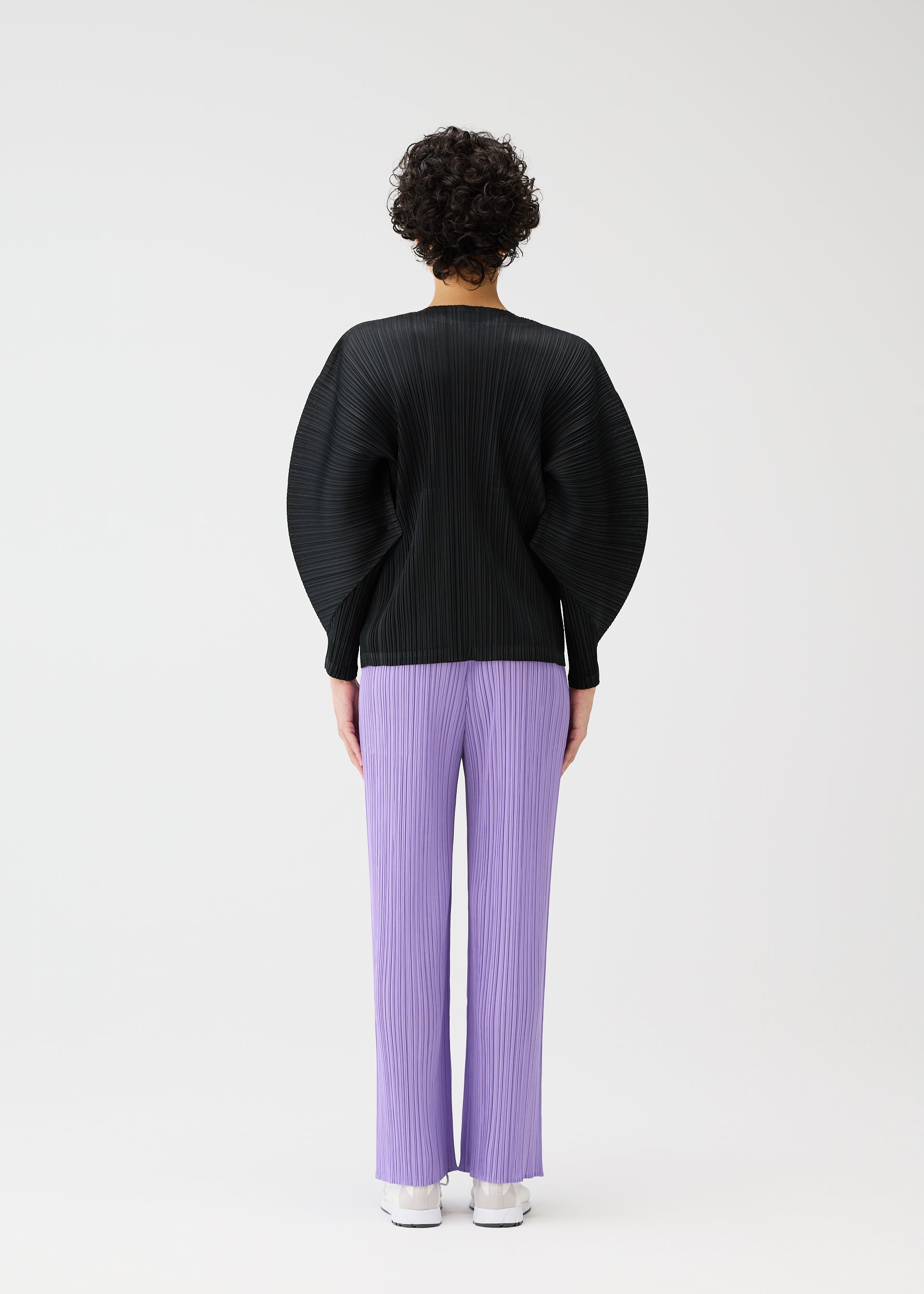 Zoomed view A person with short, curly hair stands with their back to the camera against a white background, wearing PLEATS PLEASE ISSEY MIYAKE's MONTHLY COLORS : APRIL—a black pleated oversized top with dolman sleeves and lilac pleated pants.