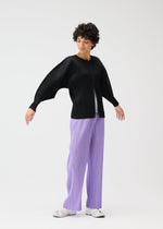 A person with short curly hair wears the MONTHLY COLORS : APRIL set by PLEATS PLEASE ISSEY MIYAKE—black pleated dolman-sleeve top and light purple pleated pants—paired with white sneakers, standing against a plain white background.