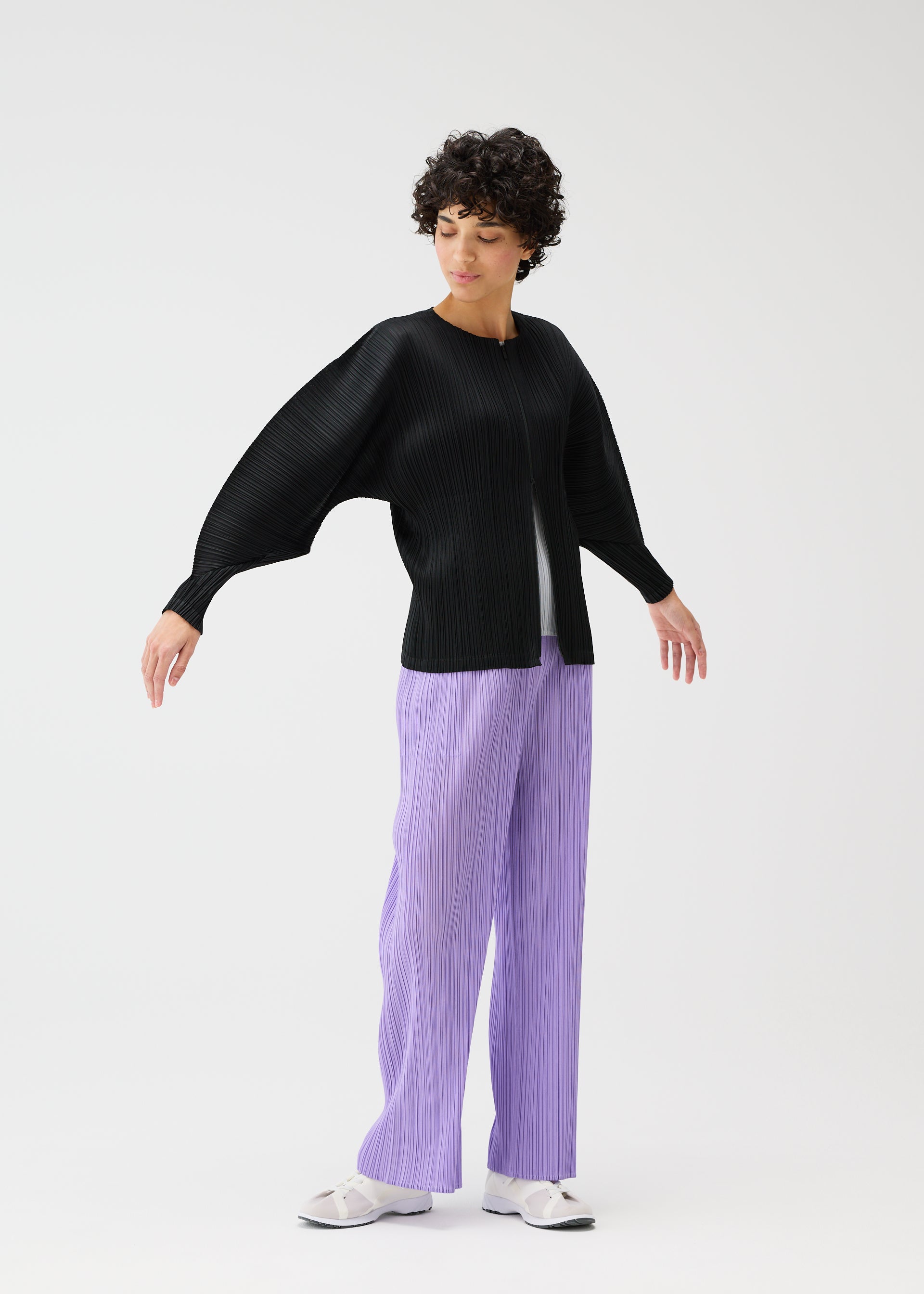 Zoomed view A person with short curly hair wears the MONTHLY COLORS : APRIL set by PLEATS PLEASE ISSEY MIYAKE—black pleated dolman-sleeve top and light purple pleated pants—paired with white sneakers, standing against a plain white background.
