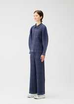 A person stands against a plain white background, wearing the COTTON DENIM textured button-up shirt and wide-leg pants from PLEATS PLEASE ISSEY MIYAKE with white shoes, facing slightly to the side and smiling softly.