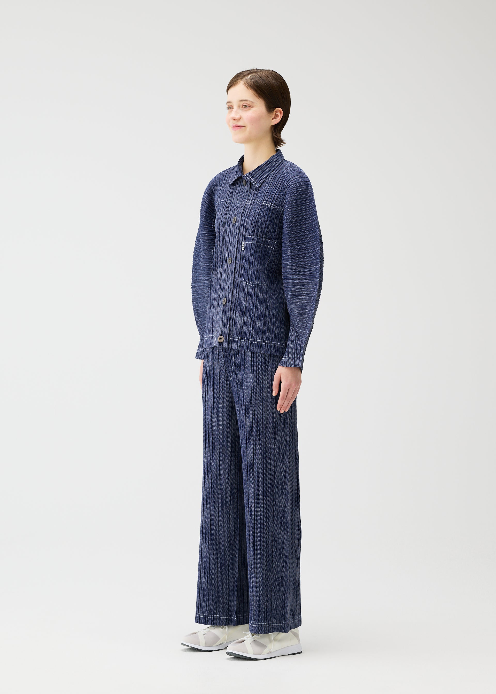 Zoomed view A person stands against a plain white background, wearing the COTTON DENIM textured button-up shirt and wide-leg pants from PLEATS PLEASE ISSEY MIYAKE with white shoes, facing slightly to the side and smiling softly.