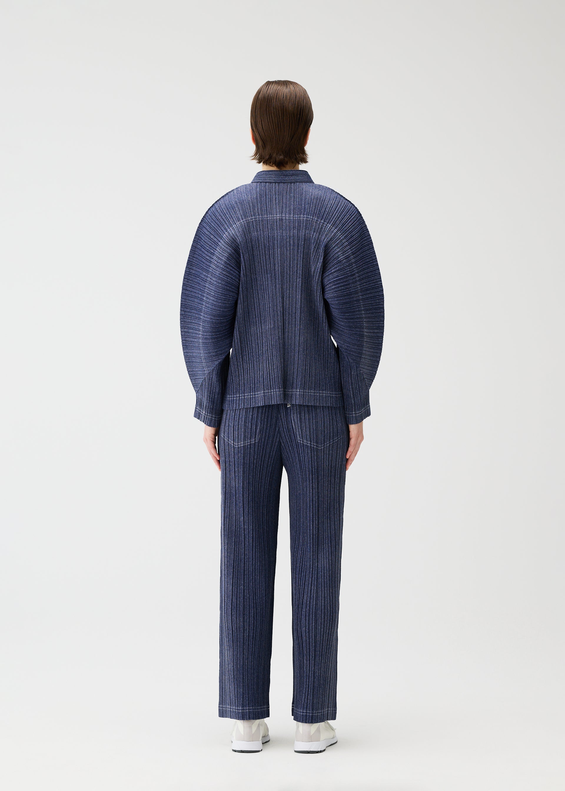 Zoomed view A person with short brown hair wears the PLEATS PLEASE ISSEY MIYAKE COTTON DENIM set—denim-inspired, collared jacket and ribbed blue pants with rounded shoulders and straight legs—paired with white shoes, against a plain light background.