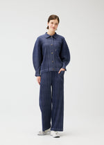 A person with short brown hair smiles against a white background, wearing the PLEATS PLEASE ISSEY MIYAKE COTTON DENIM dark blue pinstripe collared jacket and wide-leg pants with white sneakers, one hand in their pocket.