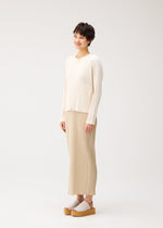 A person with short dark hair smiles in STARDUST by PLEATS PLEASE ISSEY MIYAKE, featuring a voluminous textured cardigan, beige pleated skirt, and platform sandals against a plain white background.