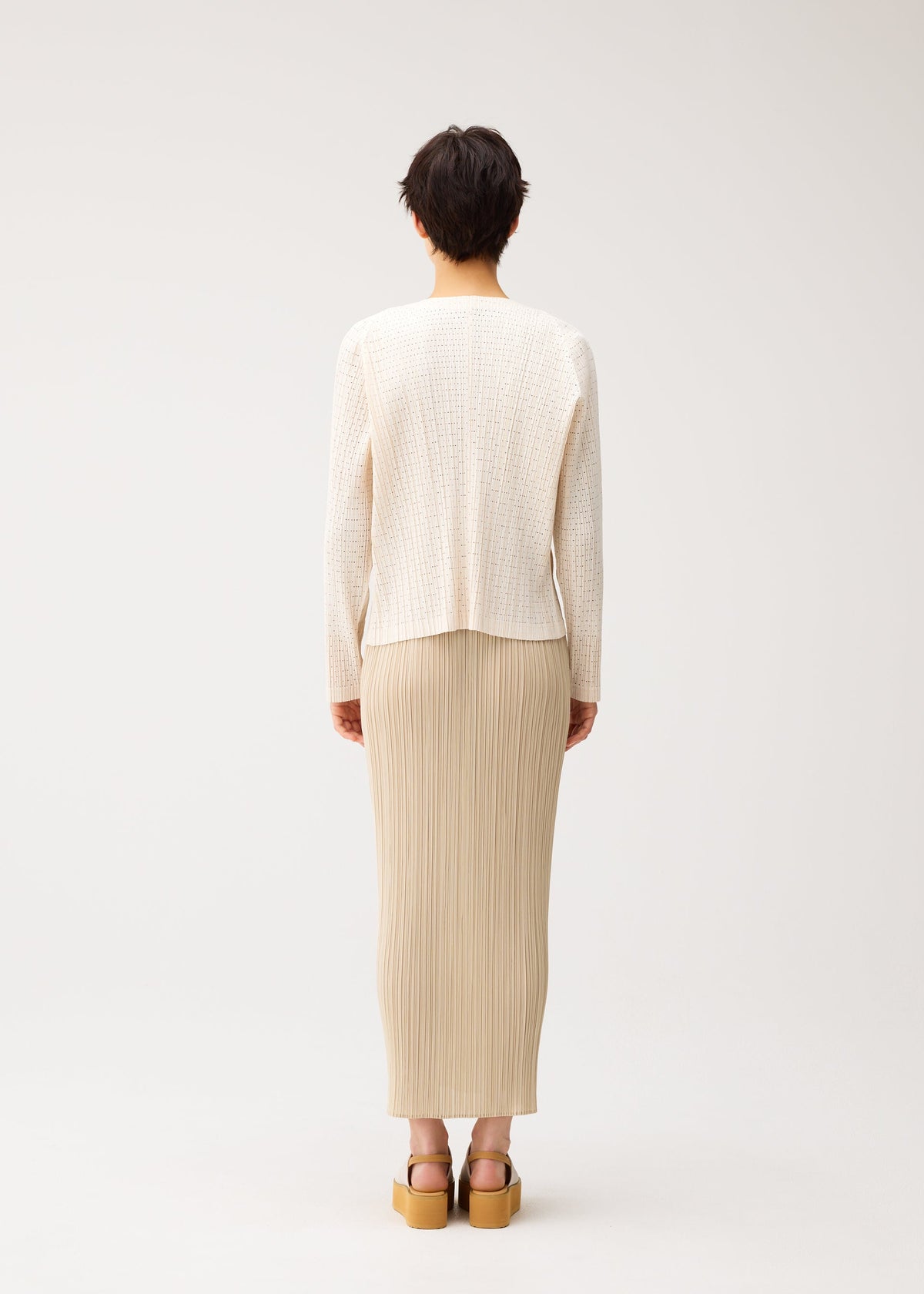 A person with short dark hair faces away, wearing the PLEATS PLEASE ISSEY MIYAKE STARDUST beige pleated midi skirt, a cream knit sweater with a voluminous fit, and tan platform sandals against a plain light background. view 8 of 11
