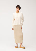 A woman with short dark hair stands against a plain background, wearing the STARDUST cardigan by PLEATS PLEASE ISSEY MIYAKE, paired with a beige pleated skirt and light platform sandals. She smiles gently, her head slightly tilted.