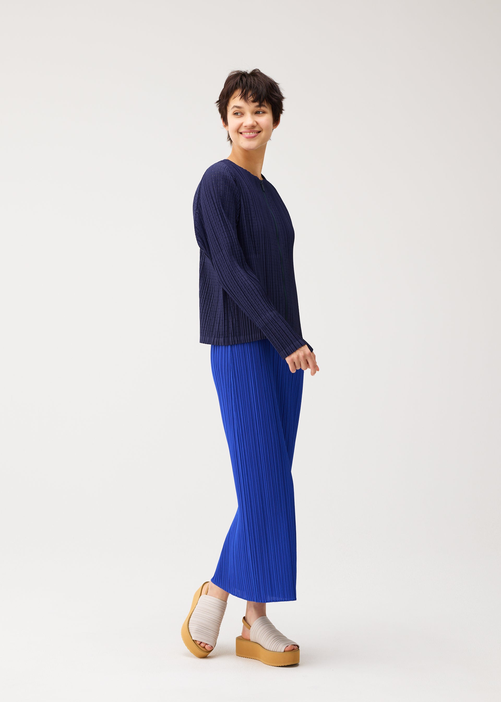 Zoomed view A person with short dark hair balances on one foot, smiling in a voluminous navy STARDUST top and bright blue pleated pants by PLEATS PLEASE ISSEY MIYAKE. They wear beige platform sandals against a plain white background.