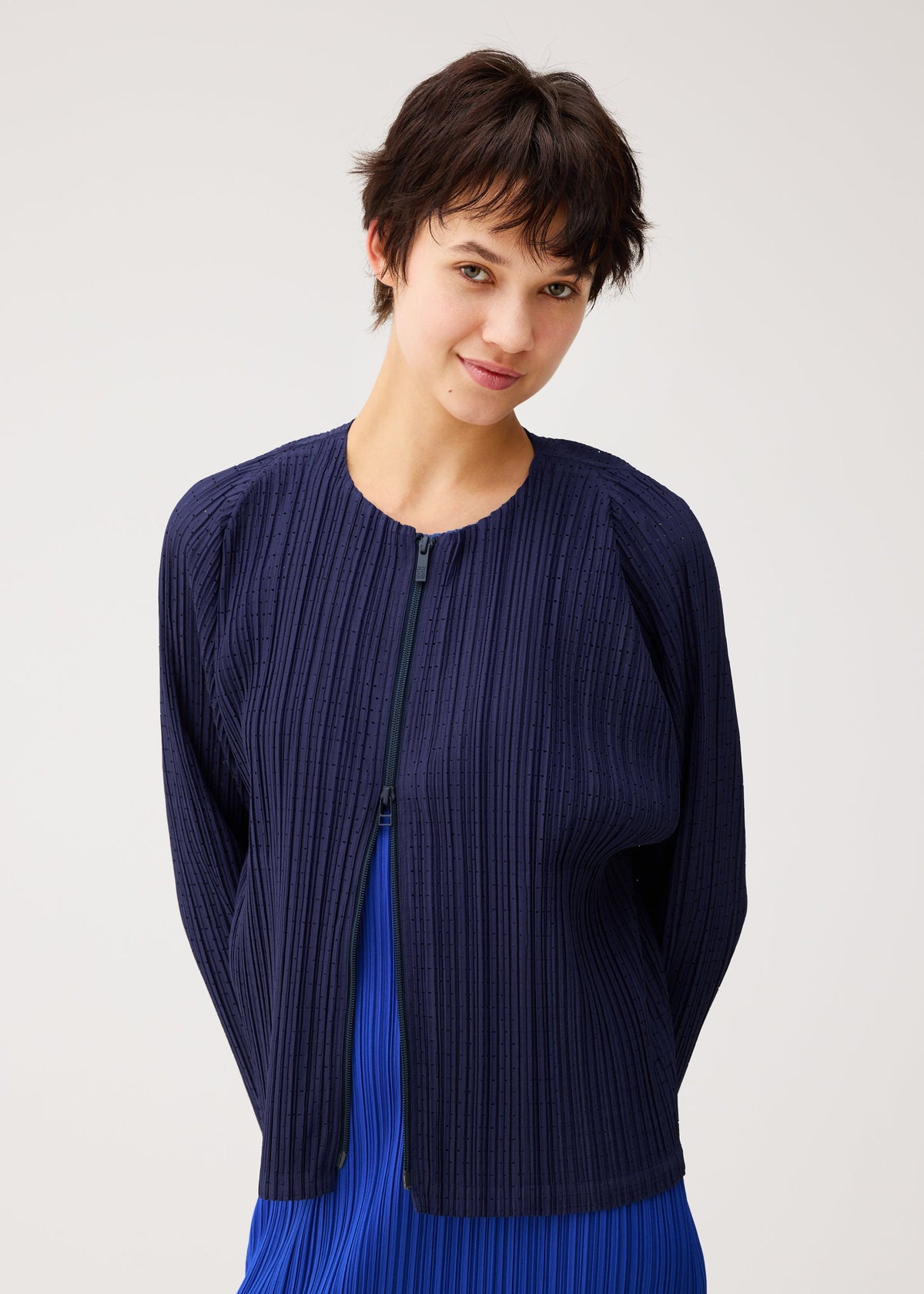 A person with short dark hair smiles gently at the camera, wearing the navy blue matte STARDUST zip-up jacket by PLEATS PLEASE ISSEY MIYAKE over a bright blue top, set against a light neutral background. view 11 of 11