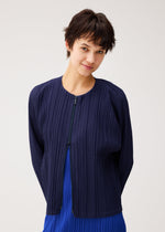 A person with short dark hair smiles gently at the camera, wearing the navy blue matte STARDUST zip-up jacket by PLEATS PLEASE ISSEY MIYAKE over a bright blue top, set against a light neutral background.