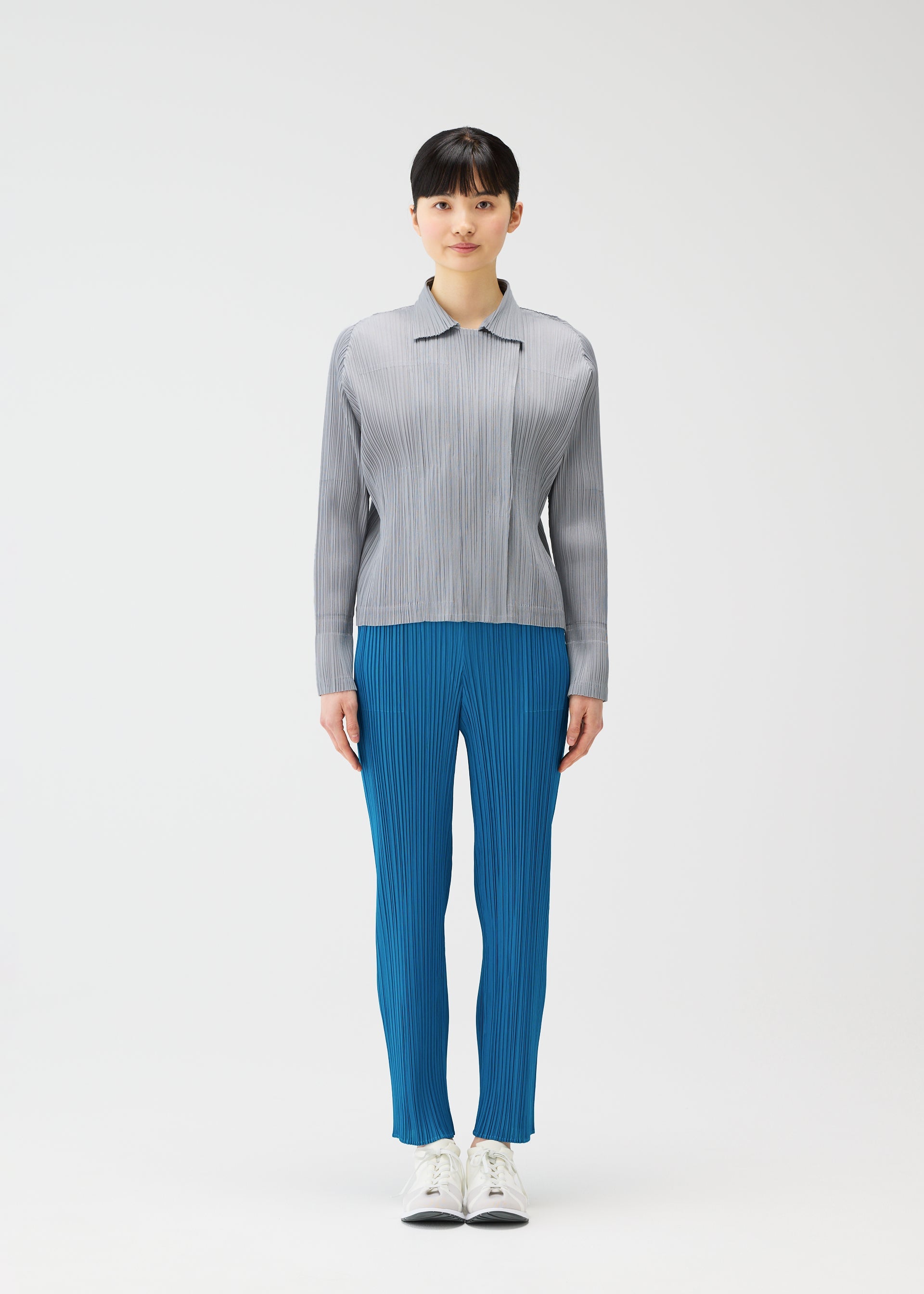 Zoomed view A person stands facing forward against a white background, wearing the light gray jacket and bright blue pleated pants from PLEATS PLEASE ISSEY MIYAKE’s MONTHLY COLORS : FEBRUARY collection, paired with white sneakers.