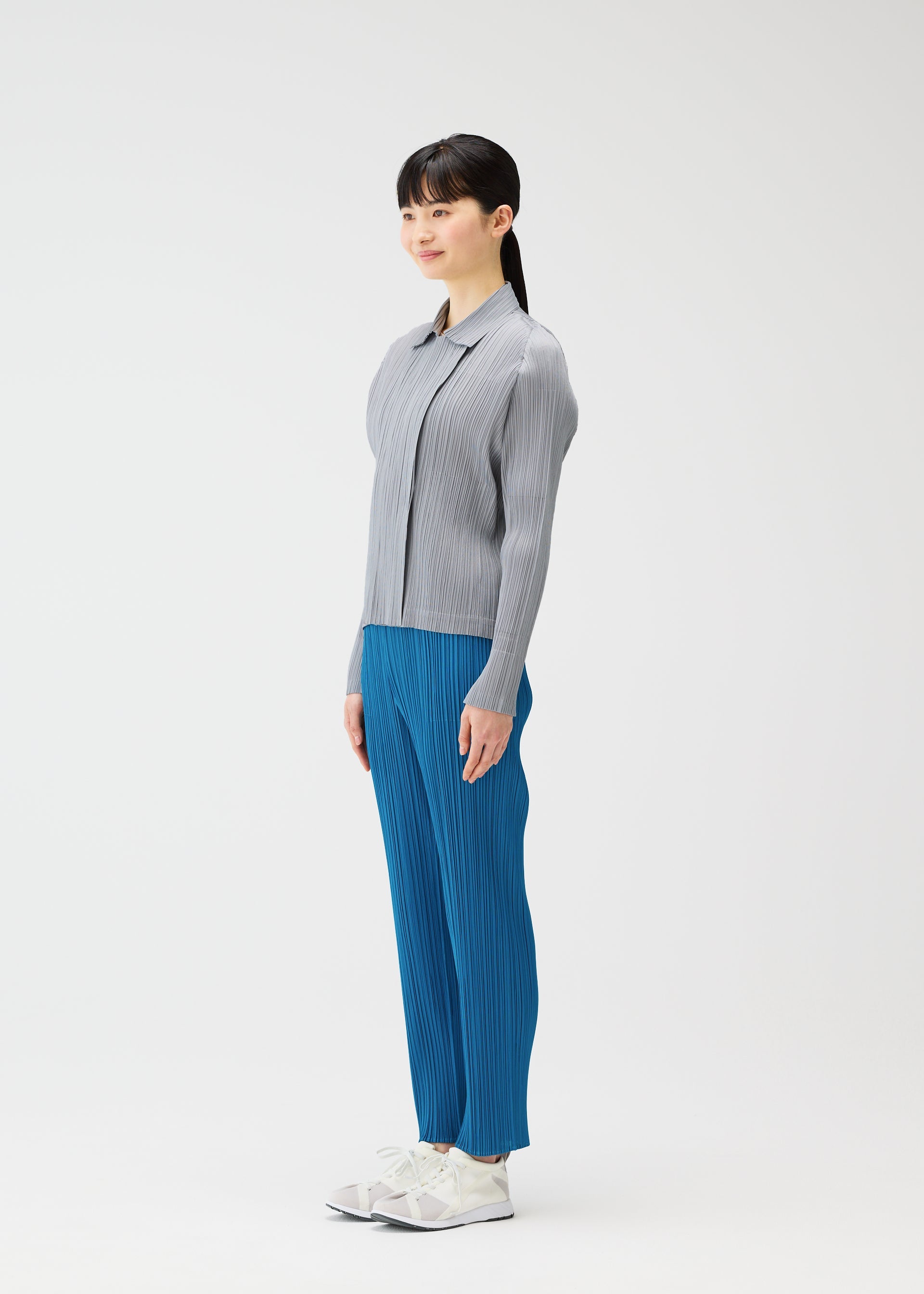 Zoomed view A woman with long dark hair stands in profile wearing the MONTHLY COLORS : FEBRUARY ribbed cardigan and blue pleated pants from PLEATS PLEASE ISSEY MIYAKE, paired with white sneakers against a plain white background.