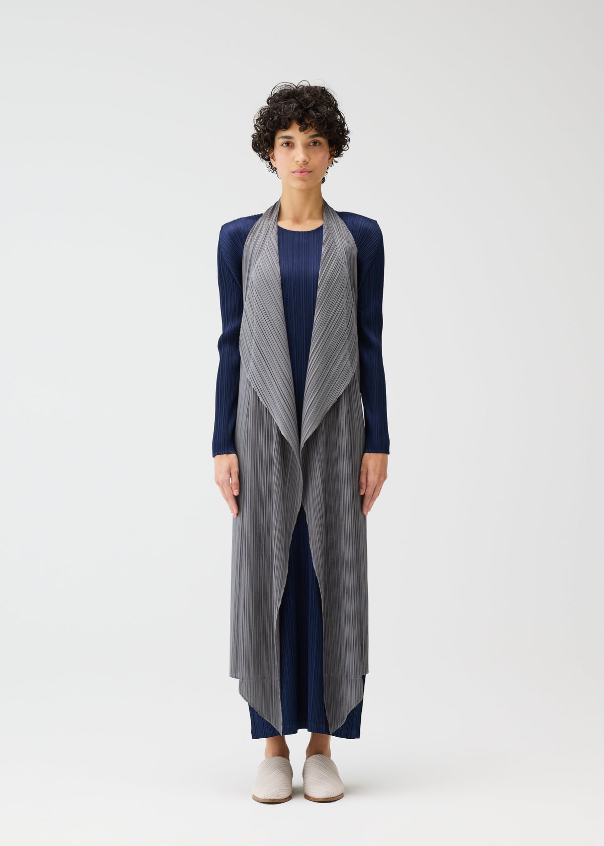A person with short curly hair stands against a plain background wearing the MONTHLY COLORS : JANUARY blue dress and textured light gray pleated vest by PLEATS PLEASE ISSEY MIYAKE, paired with light slip-on shoes. view 8 of 11