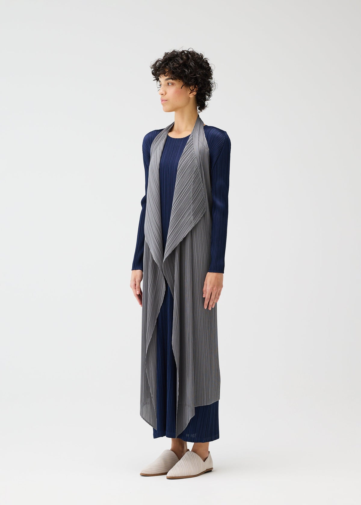 A person with short curly hair models the MONTHLY COLORS : JANUARY long navy dress and sleeveless pleated vest by PLEATS PLEASE ISSEY MIYAKE, paired with light slip-on shoes against a plain white background. view 9 of 11