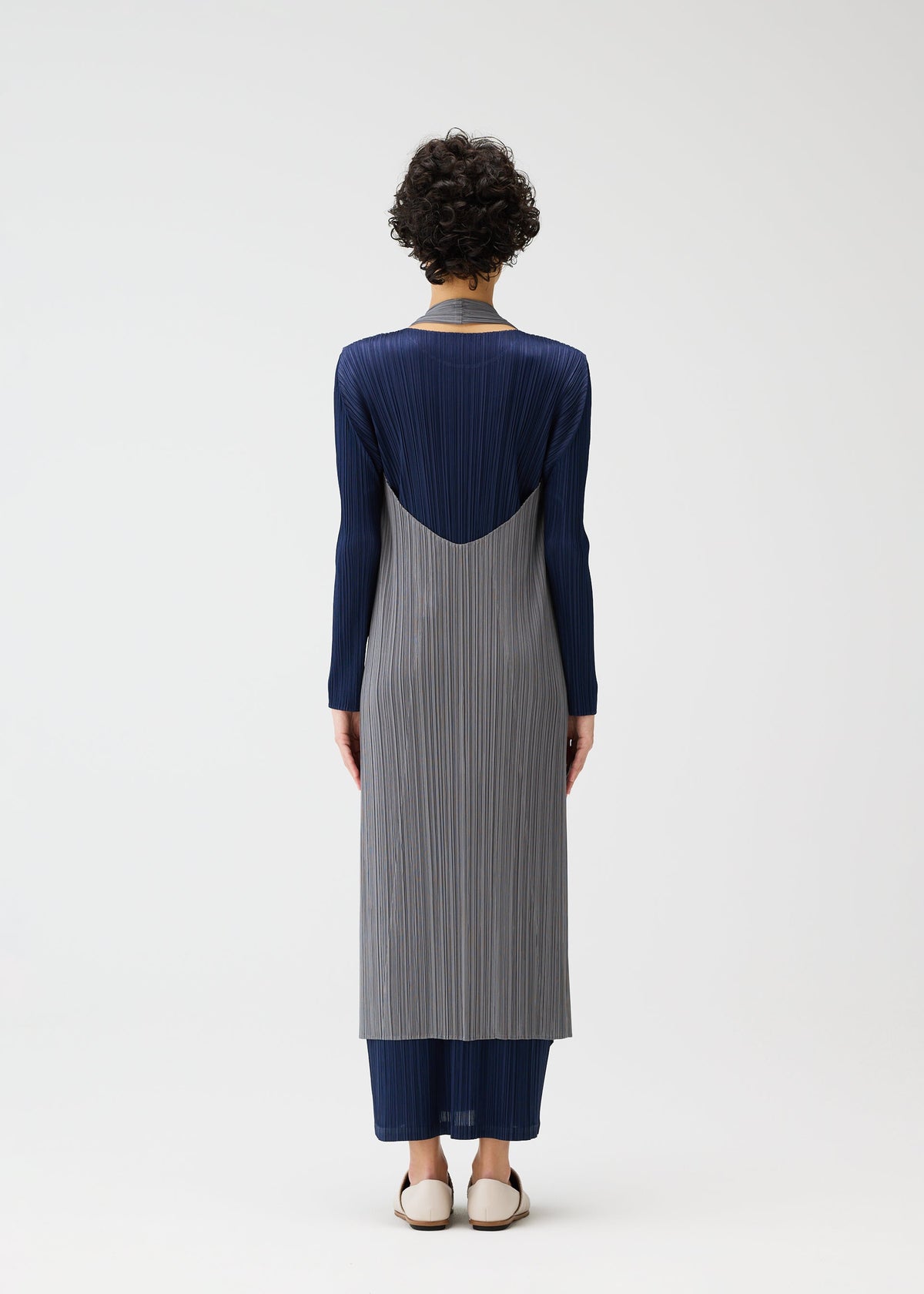 A person with short curly hair faces away, wearing PLEATS PLEASE ISSEY MIYAKE’s MONTHLY COLORS : JANUARY— a long-sleeved color-blocked dress with gray pleats, a navy layered vest, navy pants, and white slip-ons against a plain white background. view 10 of 11