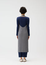 A person with short curly hair faces away, wearing PLEATS PLEASE ISSEY MIYAKE’s MONTHLY COLORS : JANUARY— a long-sleeved color-blocked dress with gray pleats, a navy layered vest, navy pants, and white slip-ons against a plain white background.