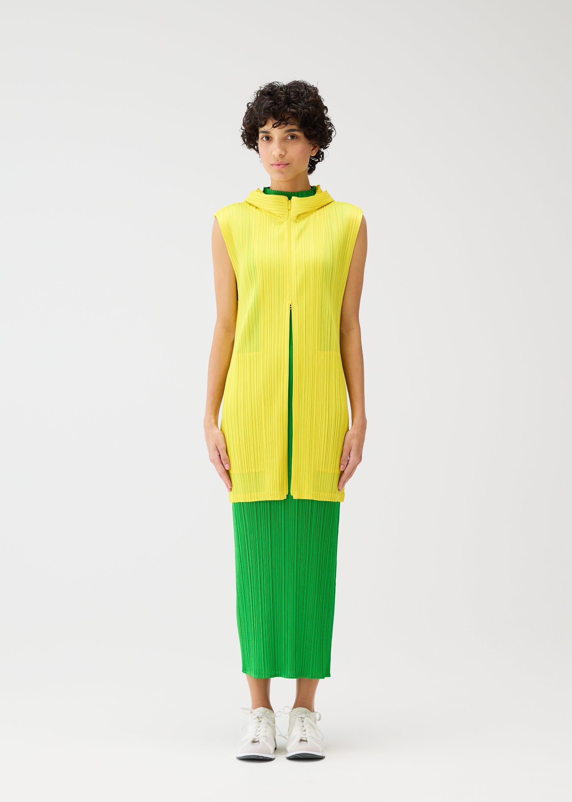 Zoomed view A person with short curly hair stands against a white background, wearing the MONTHLY COLORS : APRIL sleeveless yellow textured top and bright green skirt from PLEATS PLEASE ISSEY MIYAKE, paired with white sneakers.