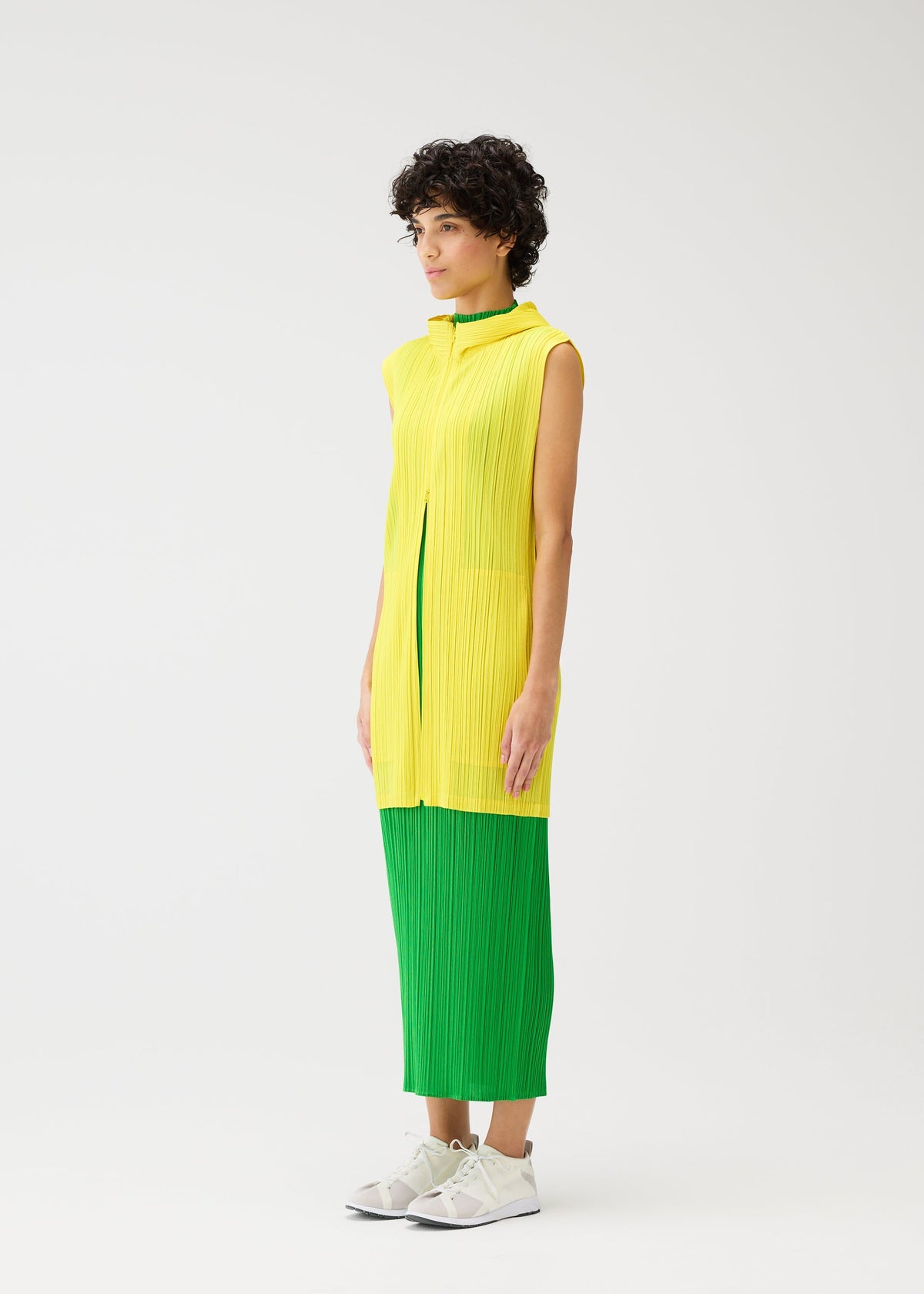A person with short curly hair wears MONTHLY COLORS : APRIL by PLEATS PLEASE ISSEY MIYAKE— a hooded long vest over a sleeveless pleated yellow top and green pleated dress, paired with white sneakers against a plain white background. view 9 of 11