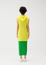 A person with short curly hair stands facing away against a white background, wearing the PLEATS PLEASE ISSEY MIYAKE MONTHLY COLORS : APRIL sleeveless yellow hooded long vest over a green skirt and white sneakers. The look features simple silhouettes.