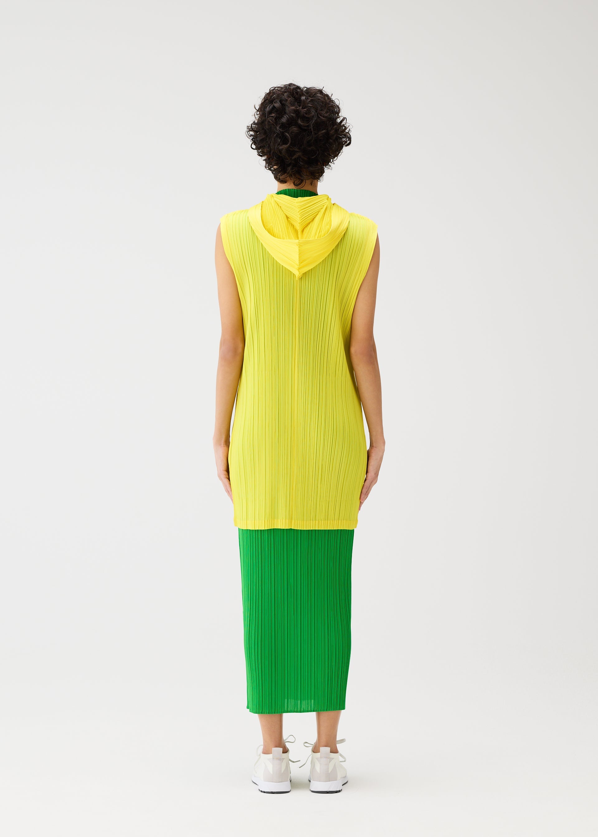 Zoomed view A person with short curly hair stands facing away against a white background, wearing the PLEATS PLEASE ISSEY MIYAKE MONTHLY COLORS : APRIL sleeveless yellow hooded long vest over a green skirt and white sneakers. The look features simple silhouettes.