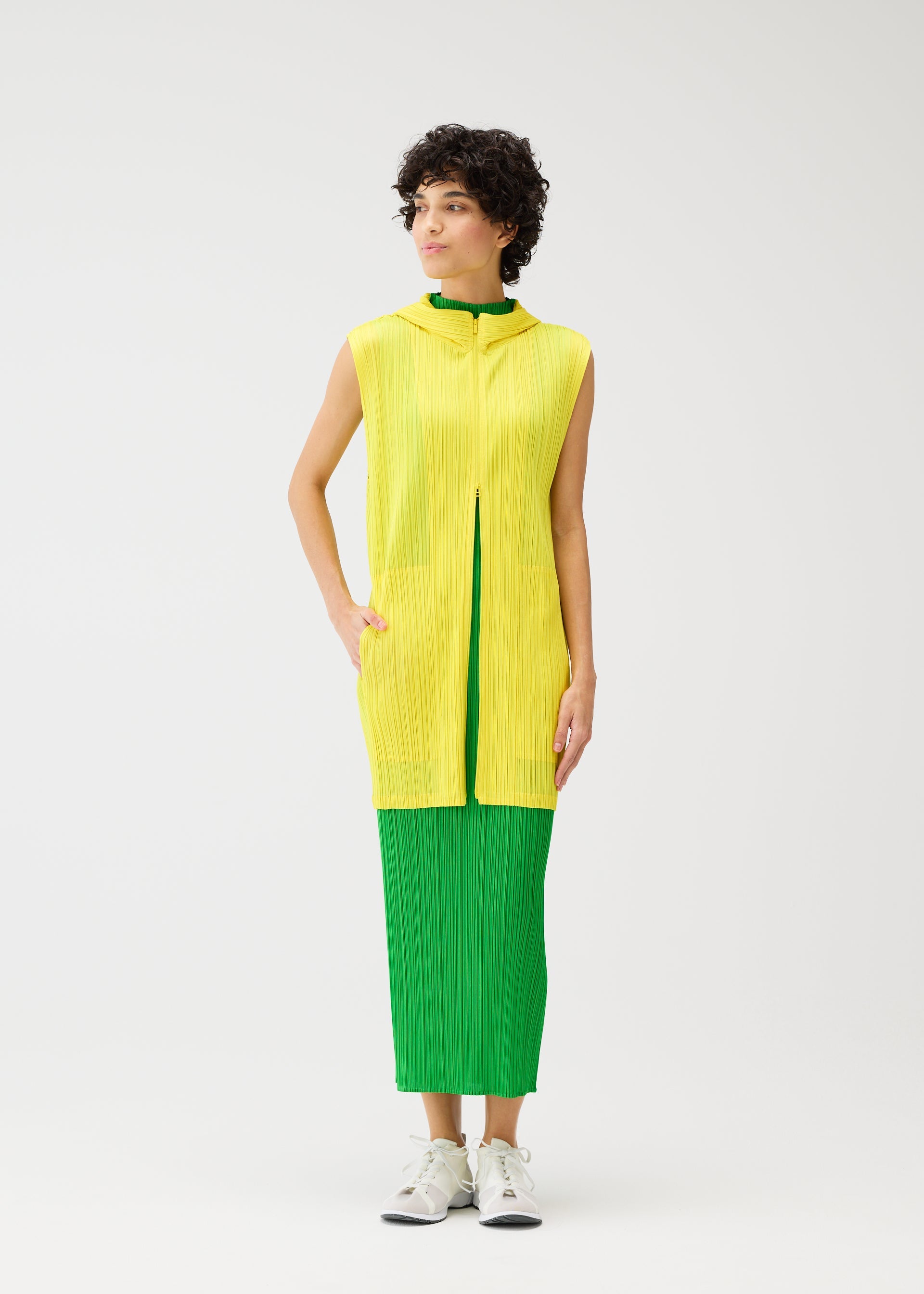 Zoomed view A person with short curly hair wears the MONTHLY COLORS : APRIL sleeveless yellow tunic by PLEATS PLEASE ISSEY MIYAKE over a green pleated dress and white sneakers, standing against a plain white background.
