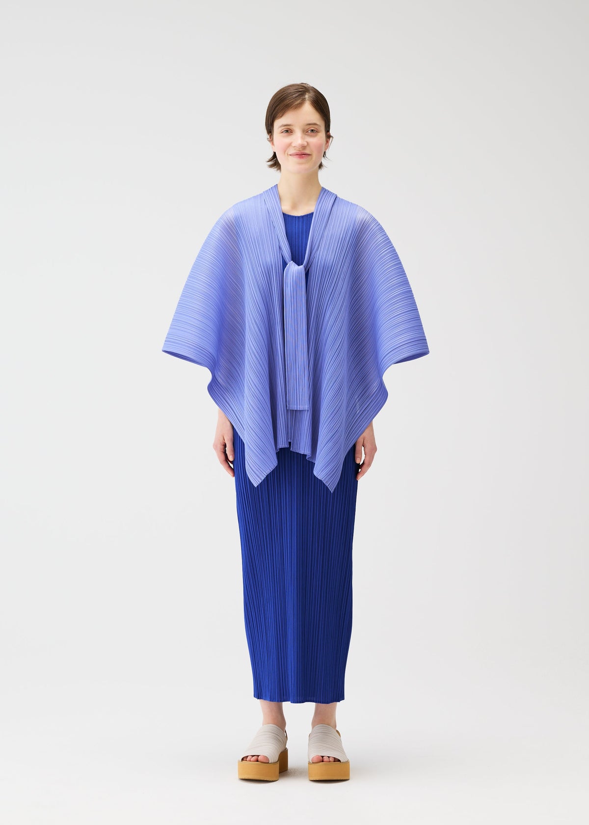 A woman with short hair and beige platform sandals stands against a white background, wearing the LUCID PLEATS dress and a 3D light blue pleated shawl by PLEATS PLEASE ISSEY MIYAKE, looking forward with a neutral expression. view 5 of 11