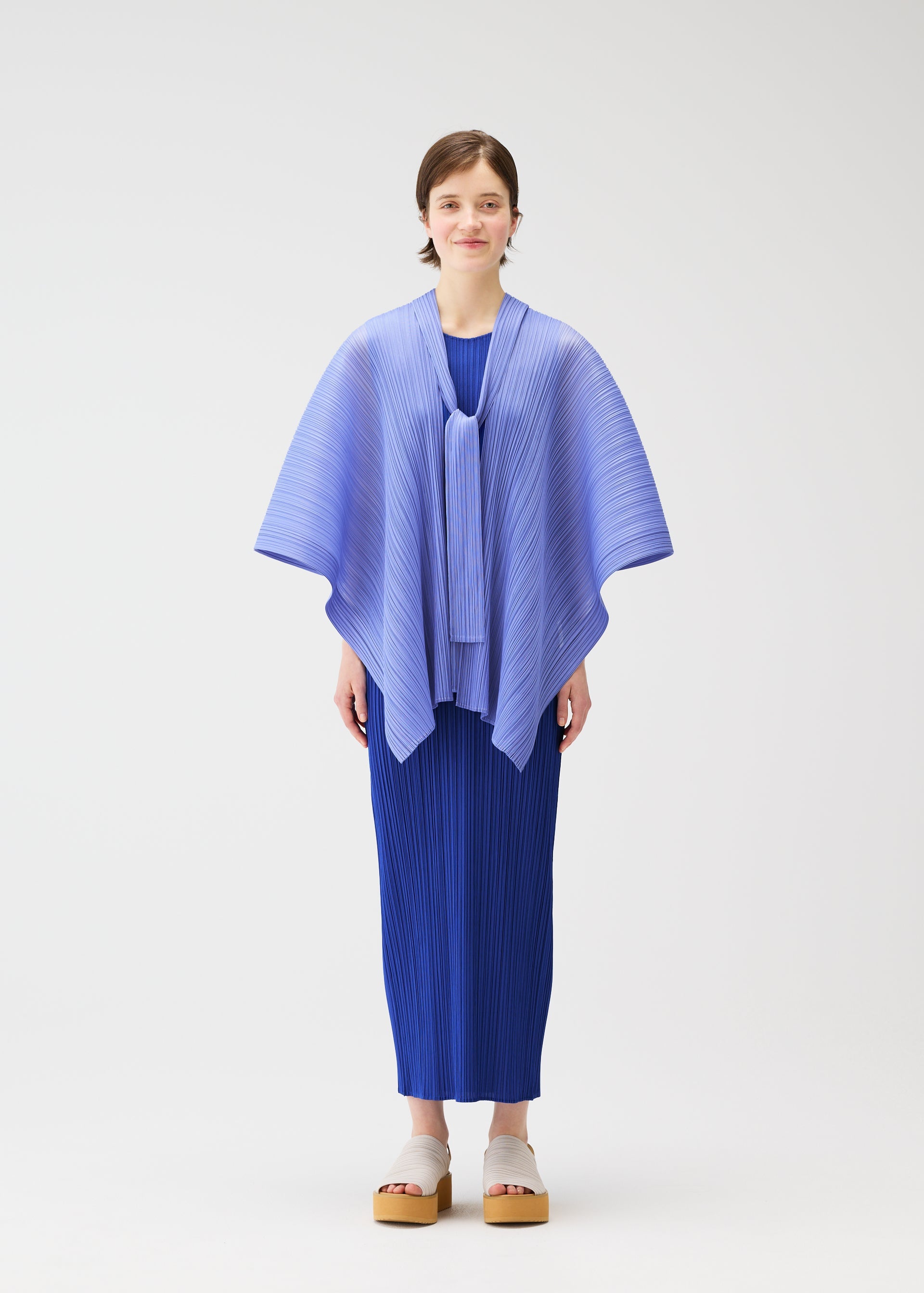 Zoomed view A woman with short hair and beige platform sandals stands against a white background, wearing the LUCID PLEATS dress and a 3D light blue pleated shawl by PLEATS PLEASE ISSEY MIYAKE, looking forward with a neutral expression.