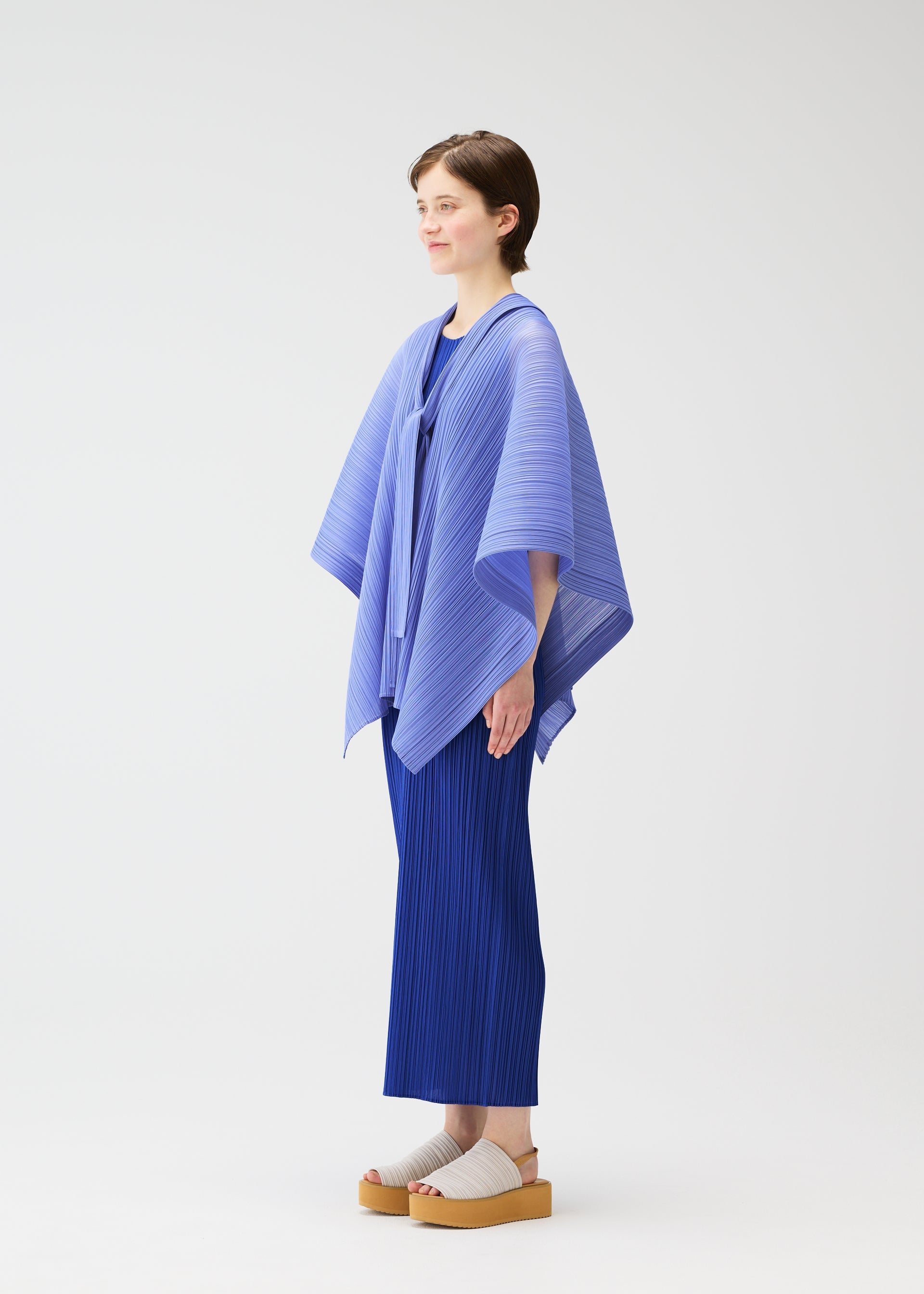 Zoomed view A person stands in profile against a white background, wearing the LUCID PLEATS set—light blue, three-dimensional cape-style top and pleated pants—from PLEATS PLEASE ISSEY MIYAKE, paired with beige platform sandals.