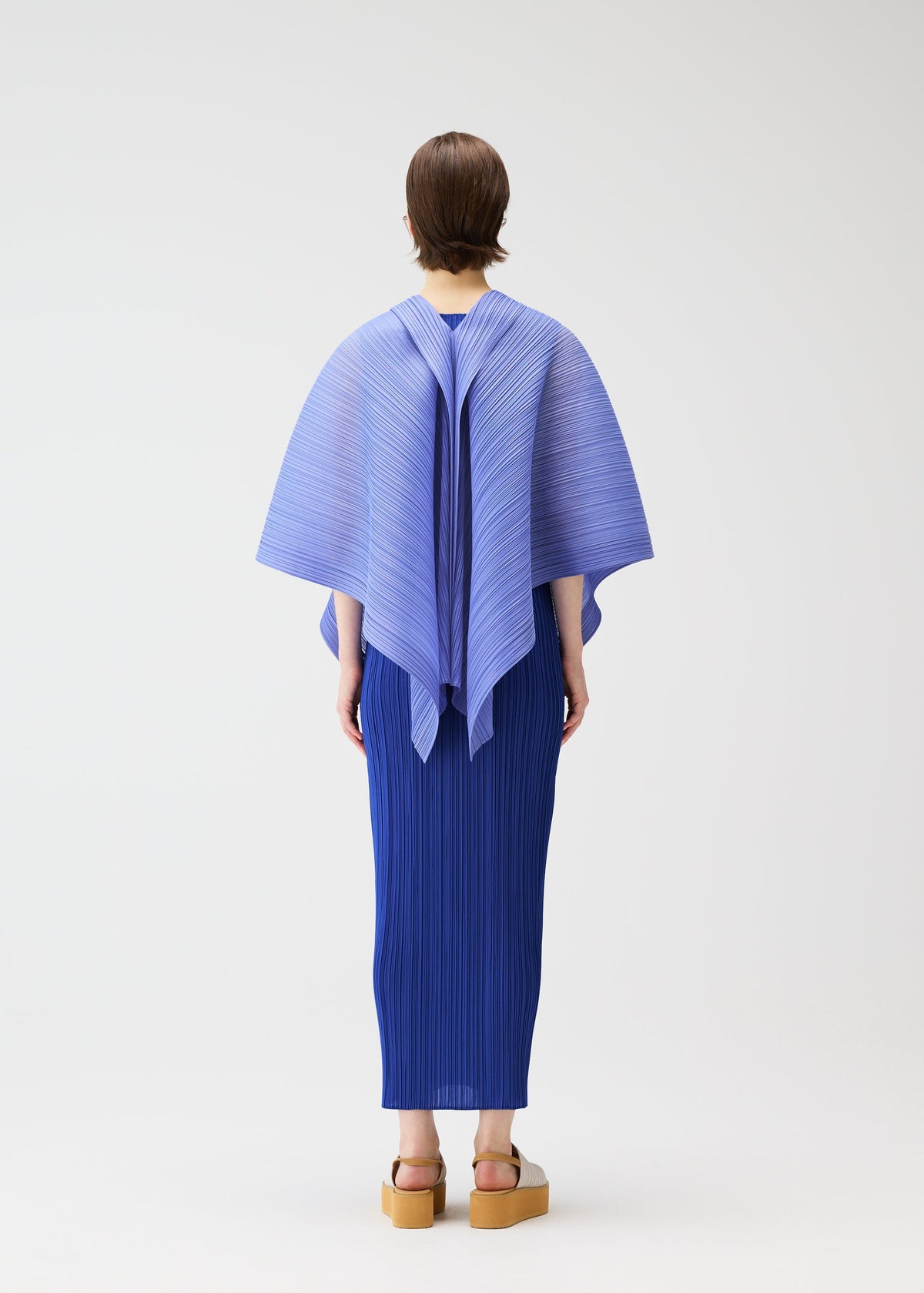 A person with short hair faces away, wearing the LUCID PLEATS blue pleated top and skirt by PLEATS PLEASE ISSEY MIYAKE, styled with tan platform sandals against a plain white background to highlight its three-dimensional design. view 7 of 11