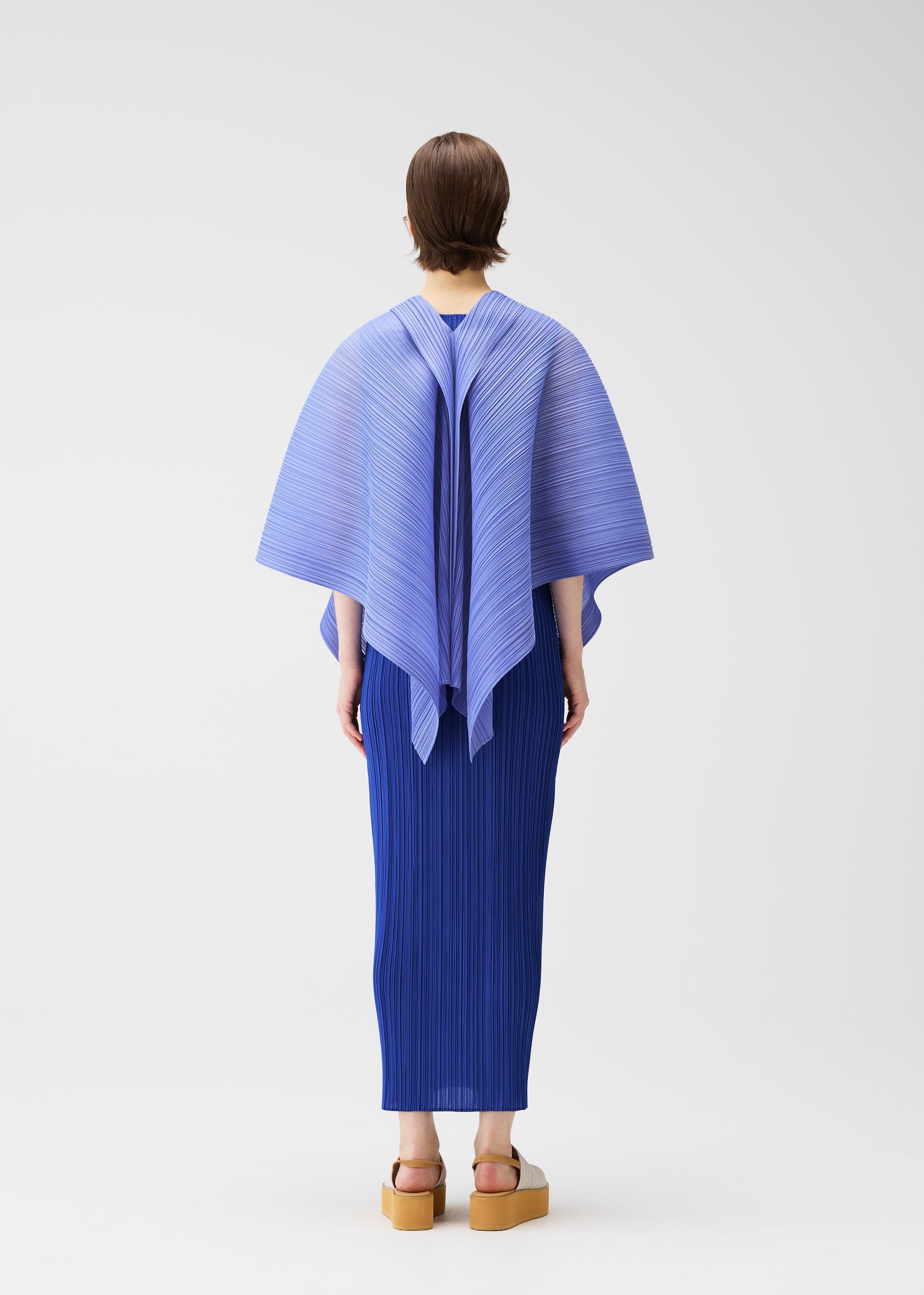 Zoomed view A person with short hair faces away, wearing the LUCID PLEATS blue pleated top and skirt by PLEATS PLEASE ISSEY MIYAKE, styled with tan platform sandals against a plain white background to highlight its three-dimensional design.