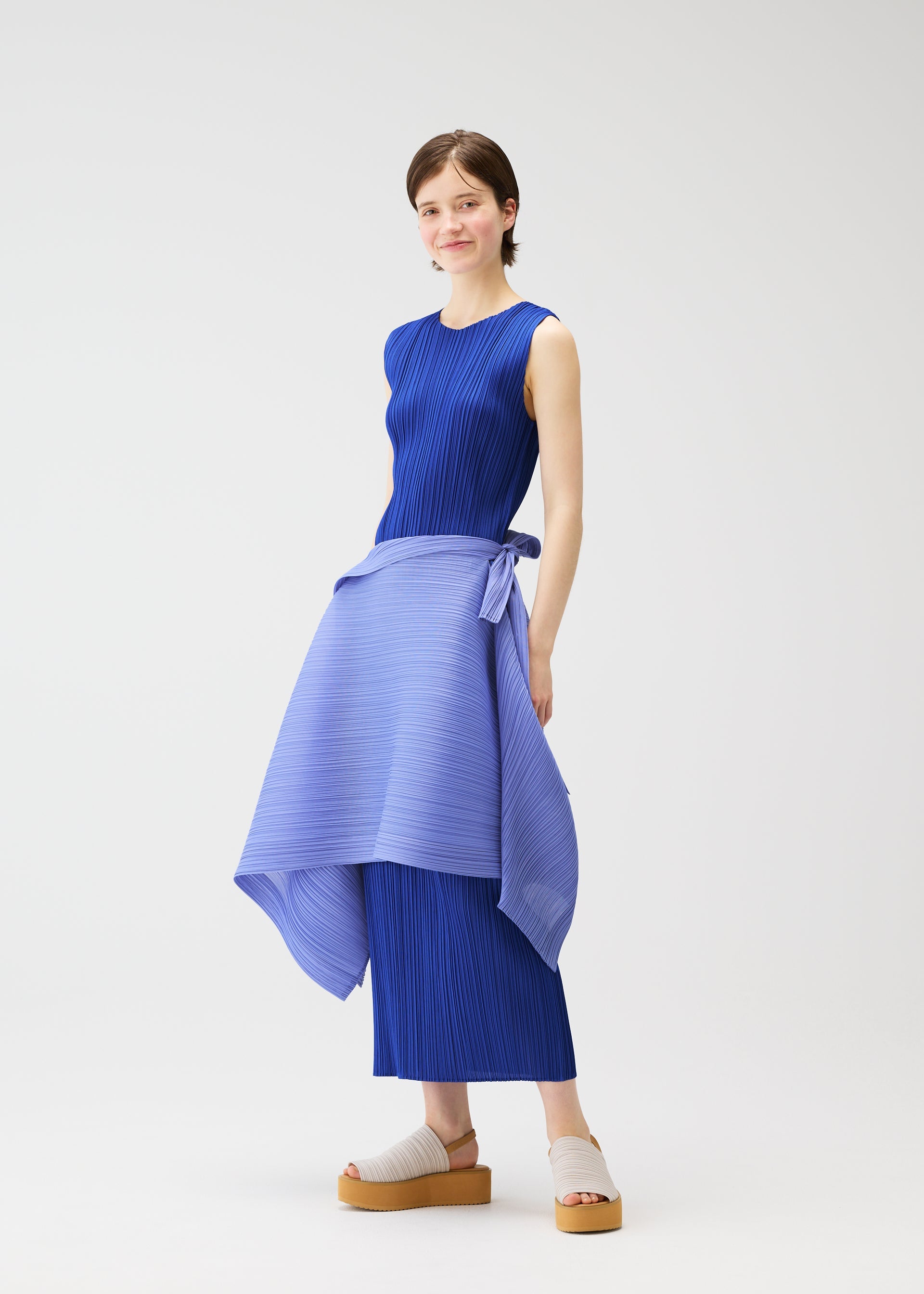 Zoomed view A woman poses against a white background wearing the PLEATS PLEASE ISSEY MIYAKE LUCID PLEATS: a sleeveless, ribbed blue dress with a pleated, light blue wrap skirt tied at the waist and platform sandals for versatile styling.