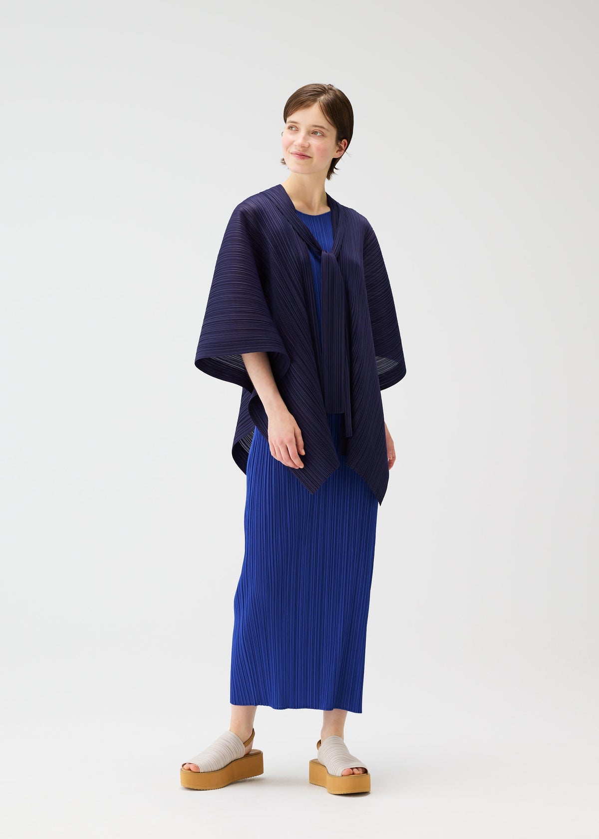 A person with short hair smiles to the side, wearing the LUCID PLEATS dress and shawl by PLEATS PLEASE ISSEY MIYAKE in blue tones, styled with beige platform sandals, against a plain white background. view 9 of 11