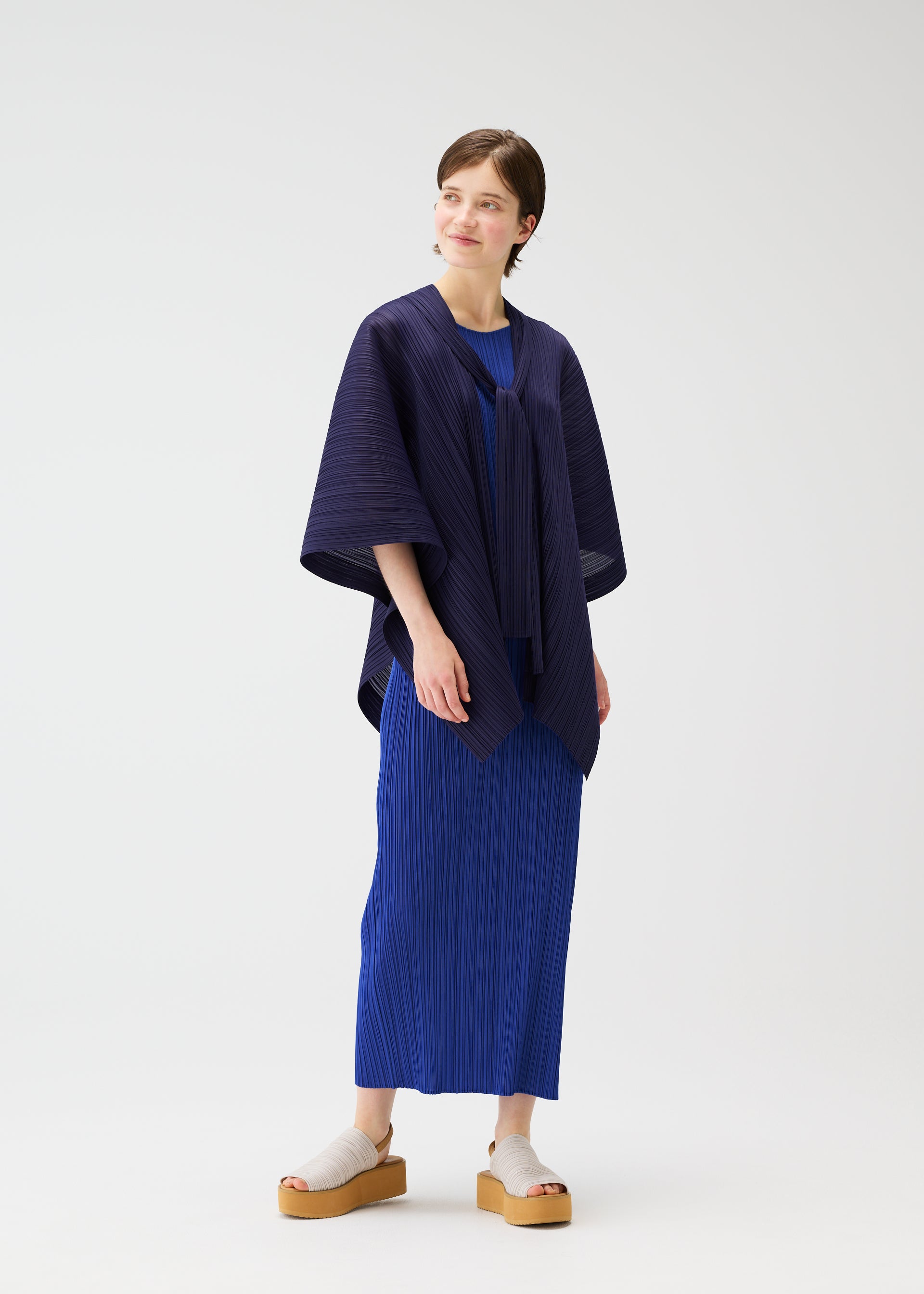 Zoomed view A person with short hair smiles to the side, wearing the LUCID PLEATS dress and shawl by PLEATS PLEASE ISSEY MIYAKE in blue tones, styled with beige platform sandals, against a plain white background.