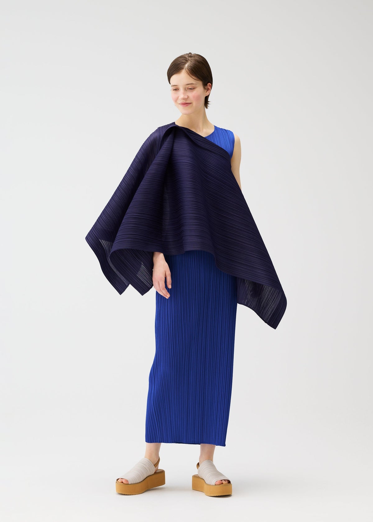 A woman models the LUCID PLEATS sleeveless blue dress by PLEATS PLEASE ISSEY MIYAKE, paired with beige platform sandals. She drapes a 3D dark blue pleated shawl over one shoulder and smiles softly against a white background. view 10 of 11