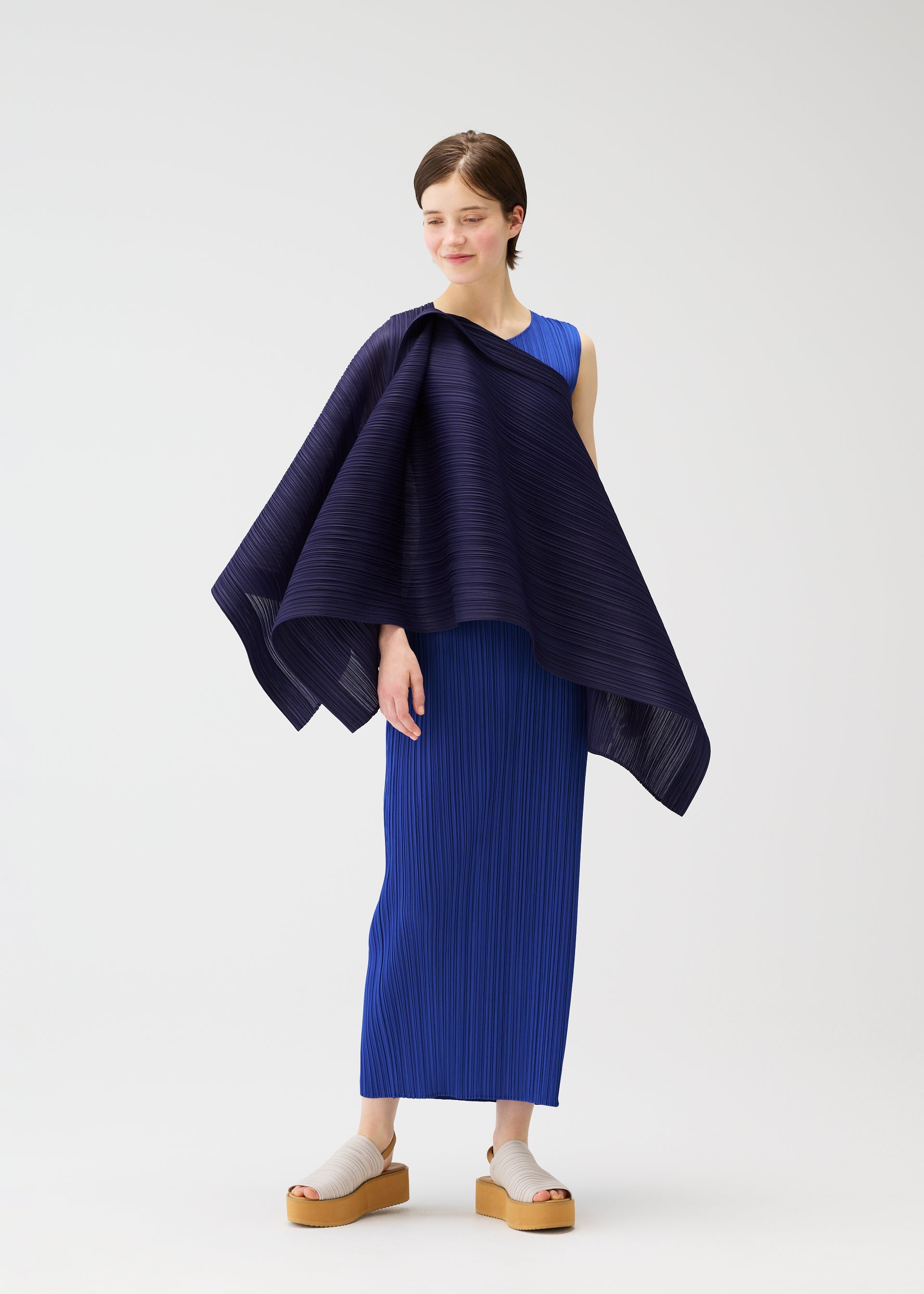 Zoomed view A woman models the LUCID PLEATS sleeveless blue dress by PLEATS PLEASE ISSEY MIYAKE, paired with beige platform sandals. She drapes a 3D dark blue pleated shawl over one shoulder and smiles softly against a white background.