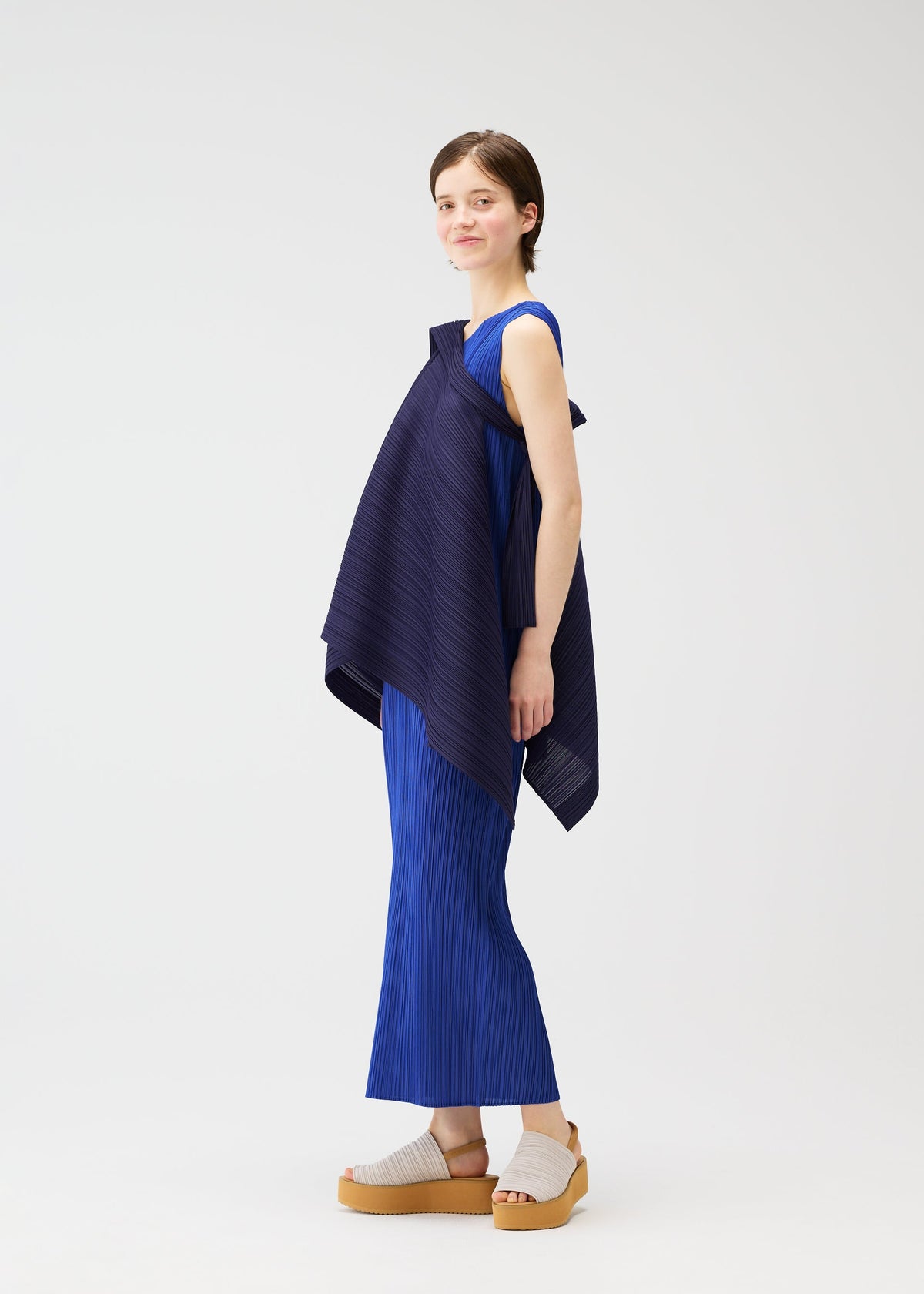 A person with short brown hair stands sideways, smiling, wearing the LUCID PLEATS set by PLEATS PLEASE ISSEY MIYAKE—a textured royal blue sleeveless top with an asymmetrical navy overlay and matching pleated pants. They pair it with beige platform sandals against a white background. view 11 of 11