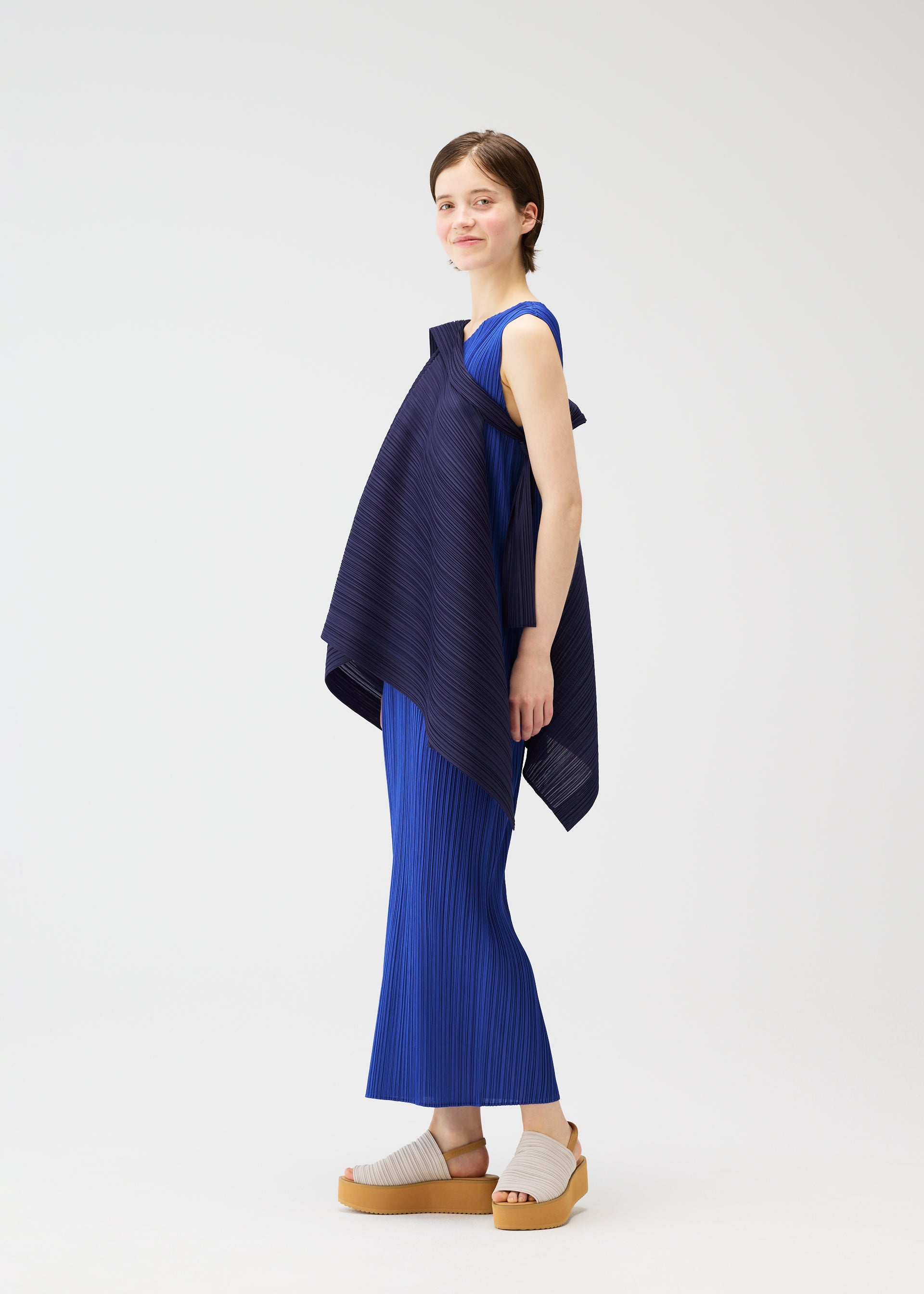 Zoomed view A person with short brown hair stands sideways, smiling, wearing the LUCID PLEATS set by PLEATS PLEASE ISSEY MIYAKE—a textured royal blue sleeveless top with an asymmetrical navy overlay and matching pleated pants. They pair it with beige platform sandals against a white background.