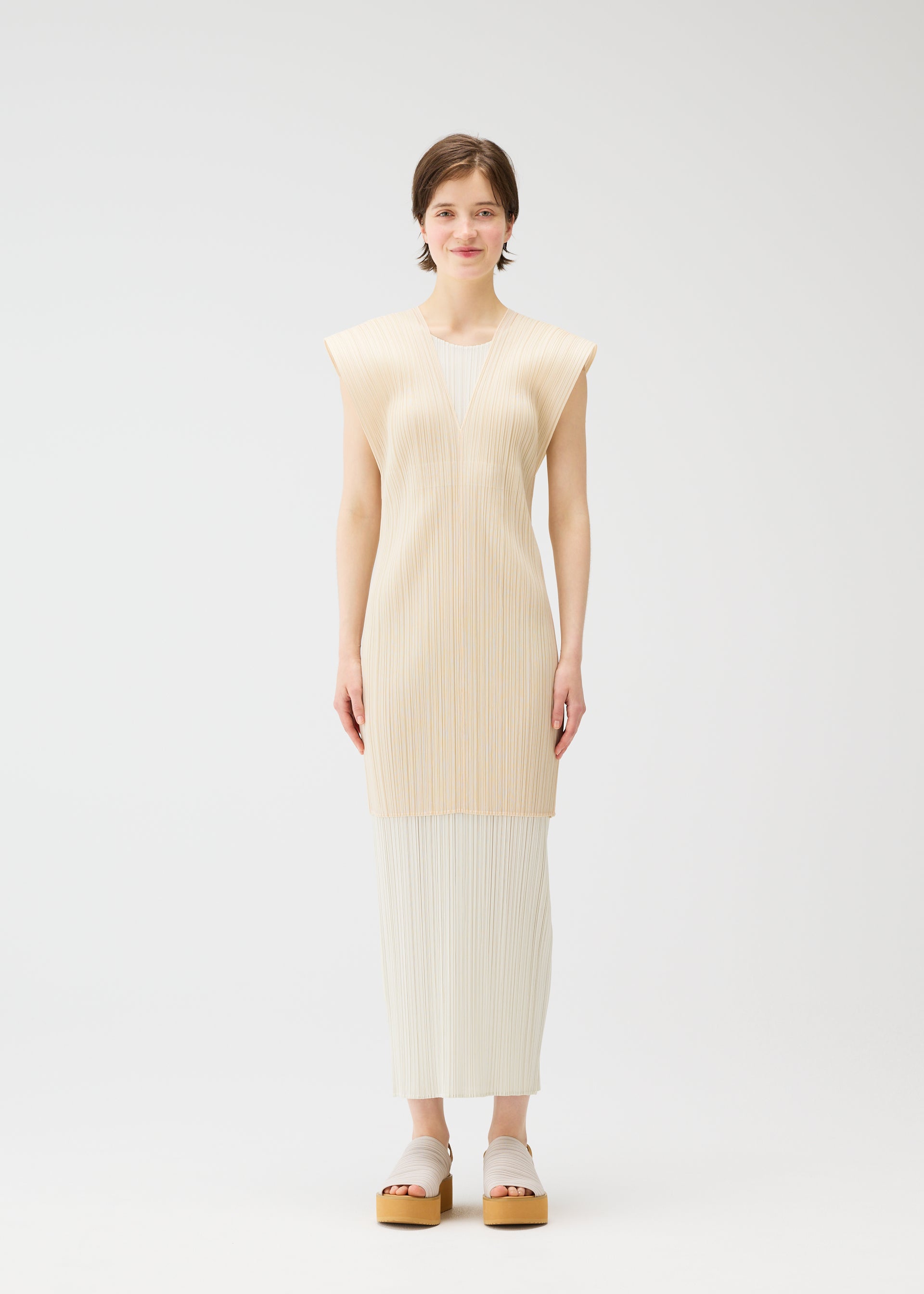 Zoomed view A woman faces forward against a plain white background, wearing the LUCID PLEATS skirt by PLEATS PLEASE ISSEY MIYAKE with a sheer long vest and white platform sandals, showcasing modern styling options.