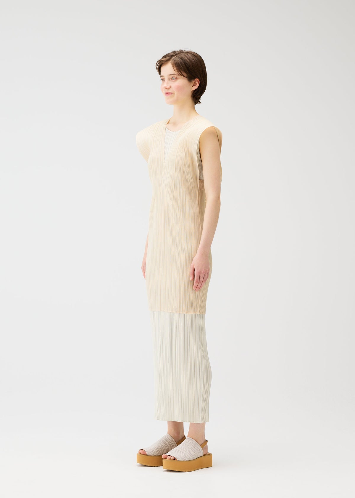 A person with short brown hair stands in a light, minimalist space, smiling gently while wearing the LUCID PLEATS collection from PLEATS PLEASE ISSEY MIYAKE—cream pleated pants, a sleeveless beige tunic, and a sheer long vest. view 6 of 9